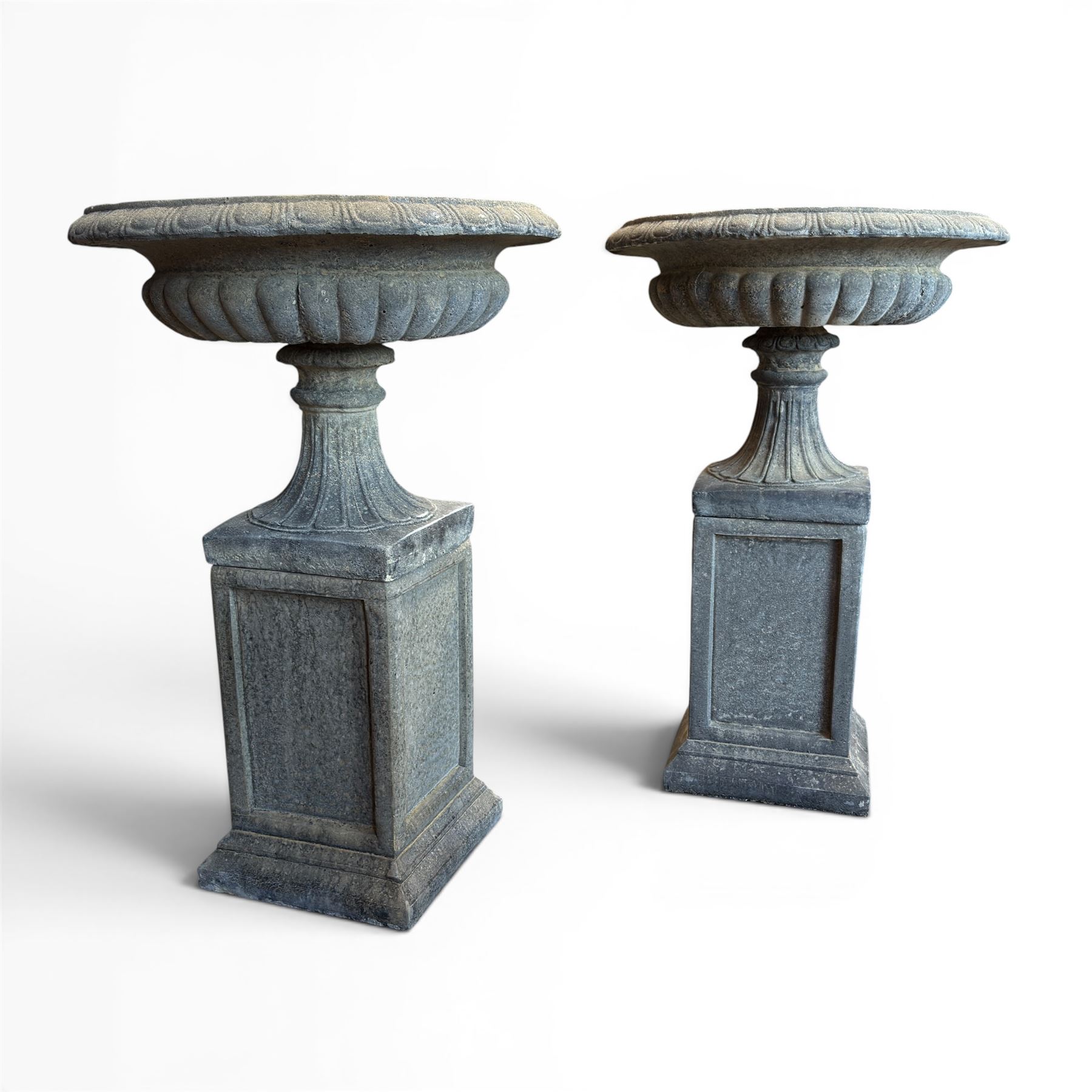 Pair of cast stone garden urns of kylix form on pedestals, the broad egg-and-dart moulded rim over a shallow gadrooned underbelly, with waisted tapering base, raised on matching plinth bases with faux panelled facia