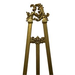 Victorian design gilt floor standing picture easel, the C-scroll and leaf decorated pediment over the moulded frame with upper extending flower head decoration, the apron and terminals in the form of S-scrolled foliate and central fanned leaf cartouche, with adjustable picture rest  