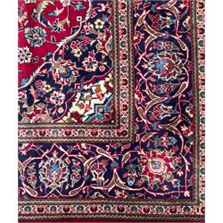 Central Persian Kashan hand-knotted carpet, the deep red ground with a central lozenge medallion in ivory and navy, surrounded by dense scrolling floral and vine motifs, navy spandrels and a wide border with stylised palmettes and flowerheads enclosed by multiple guard stripes