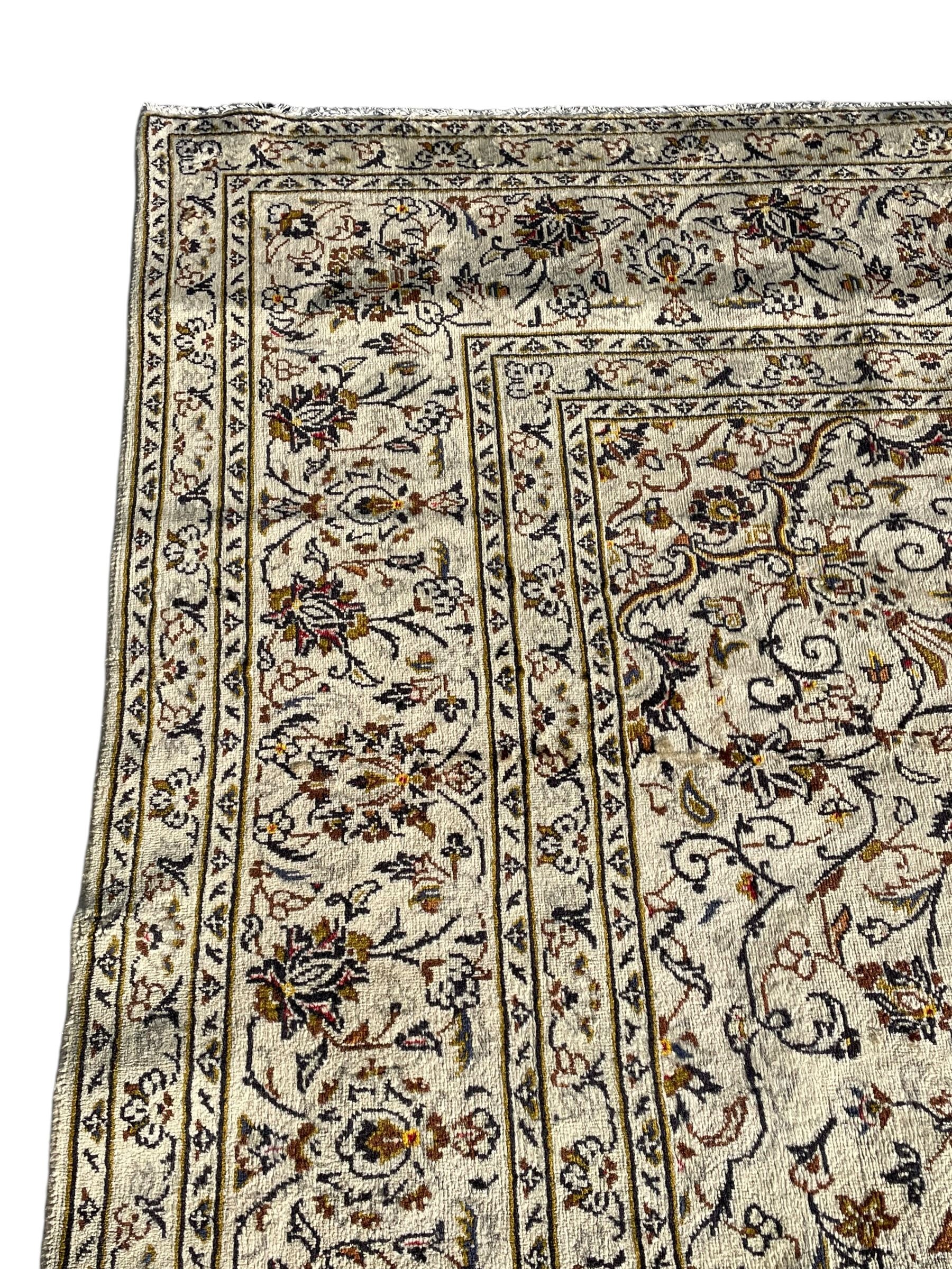 Persian Kashan ivory ground carpet, central medallion decorated with sixteen arranged palmette motifs, within a field decorated with swirling leafy branches and stylised plant motifs, the main border decorated with matching swirling pattern, within floral pattern guard stripes 