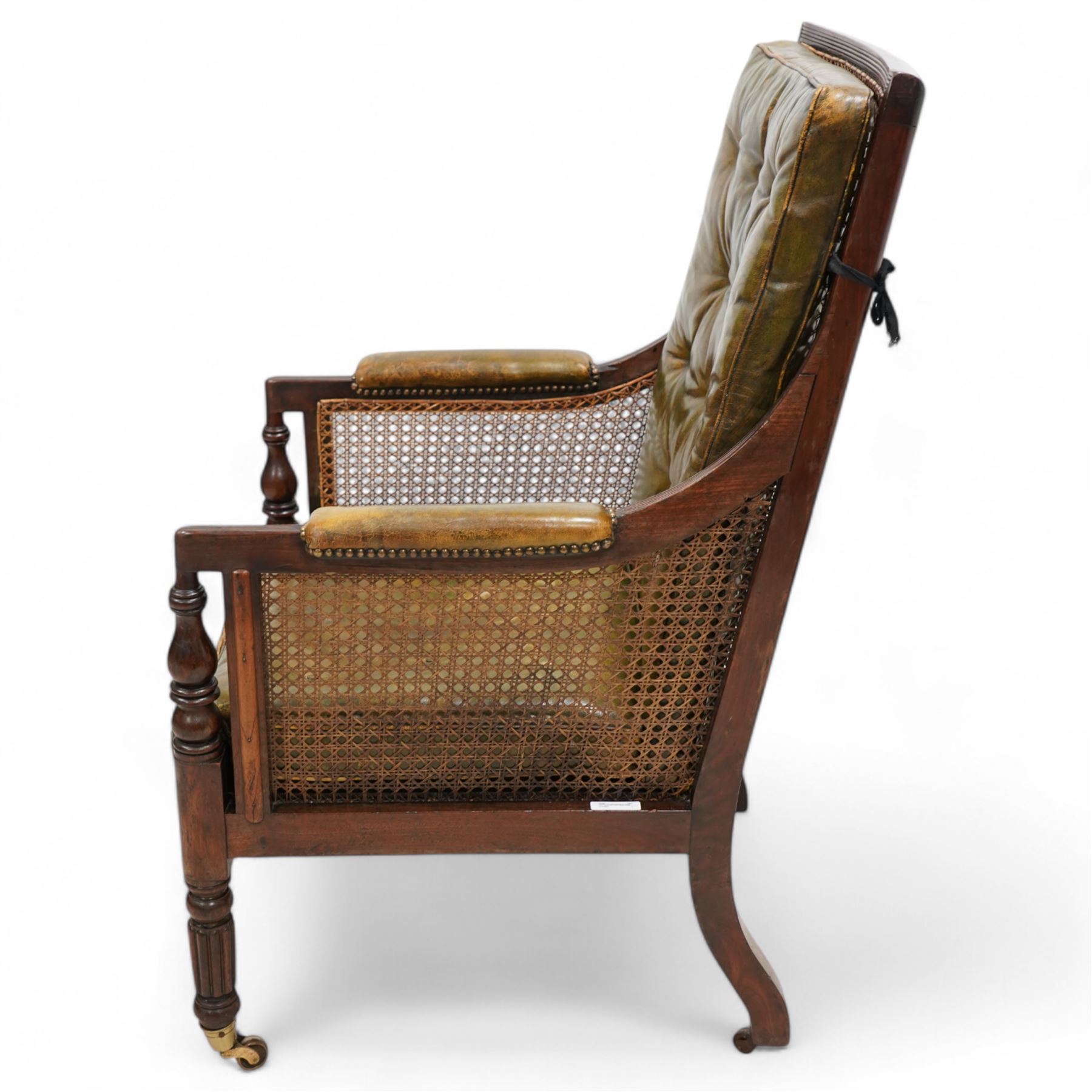 George IV mahogany framed library reading chair, reed moulded frame with cane work seat, back and sides, with loose cushions upholstered in green buttoned leather, upholstered arm rests, turned arm supports terminating to turned and reed carved feet, on castors 