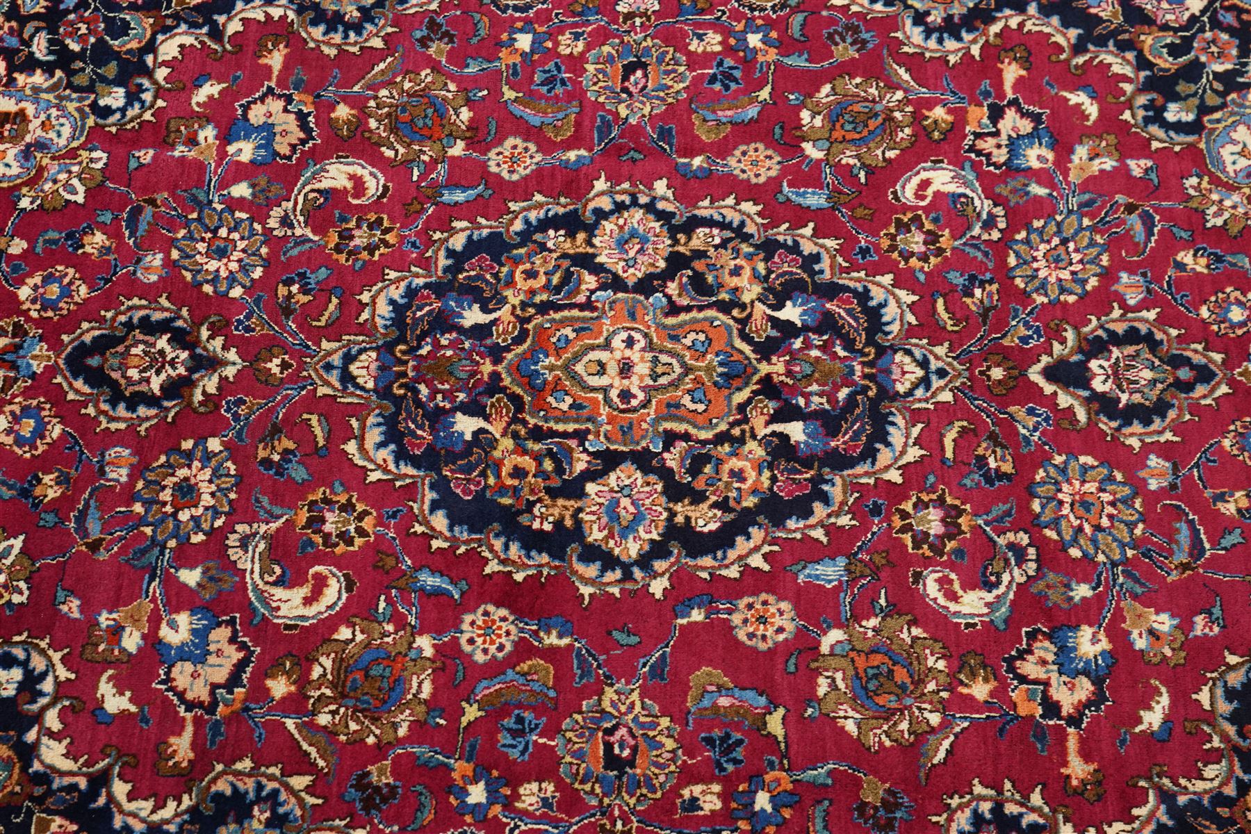Central Persian Kashan hand-knotted carpet, rose red field, with central shaped lobed medallion issuing an all-over scrolling design of palmettes, floral sprays and arabesques, within conforming spandrels, enclosed by a broad indigo herati type palmette border framed by multiple guard stripes, in a palette of deep red, midnight blue and ivory tones, with cotton fringe ends, and Kashan signature 