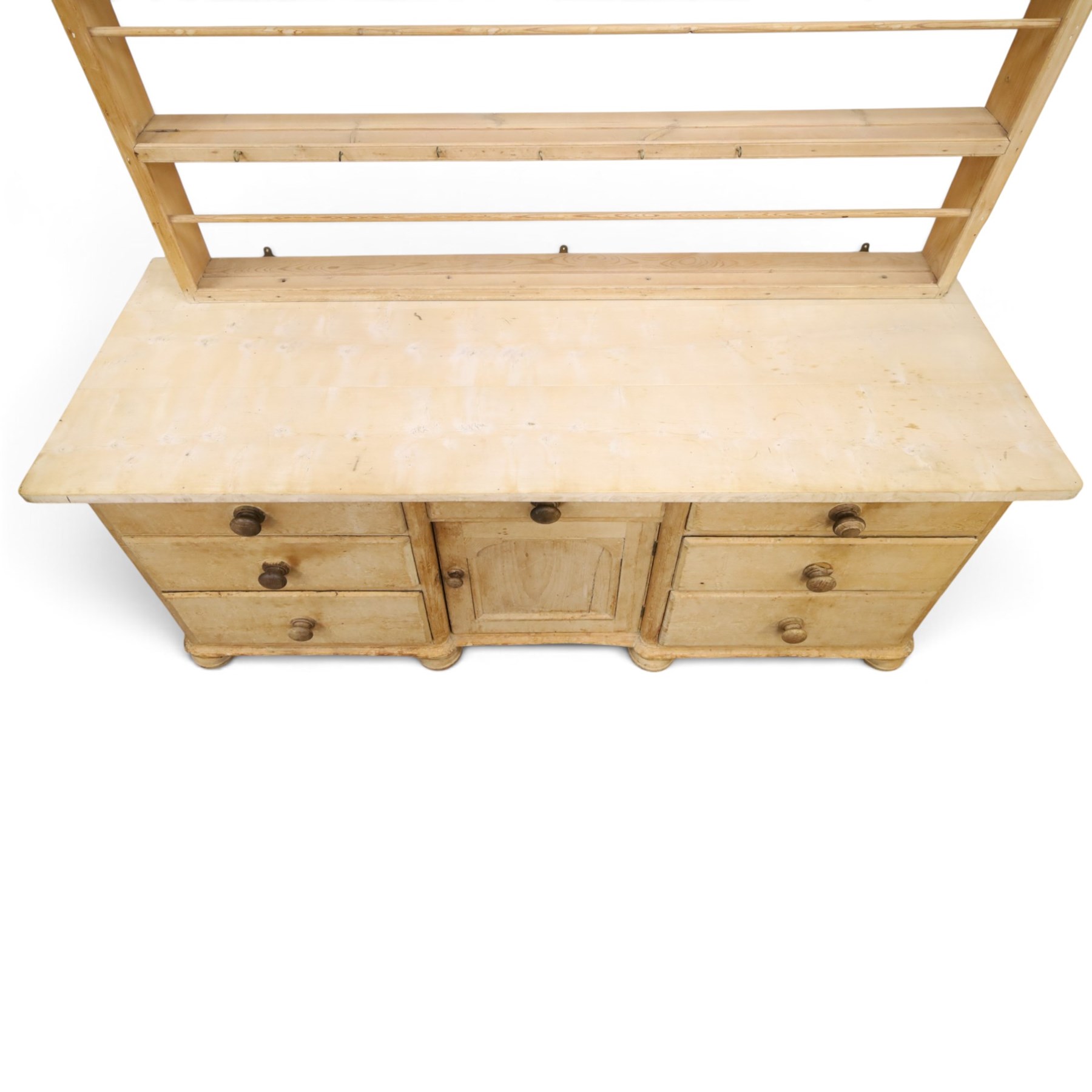 Victorian stripped pine dresser base with plate rack, the four-tier plate rack with supporting rails and hooks, the inverted-breakfront base fitted with seven assorted graduating drawers surrounding a recessed cupboard, on compressed bun feet with castors
