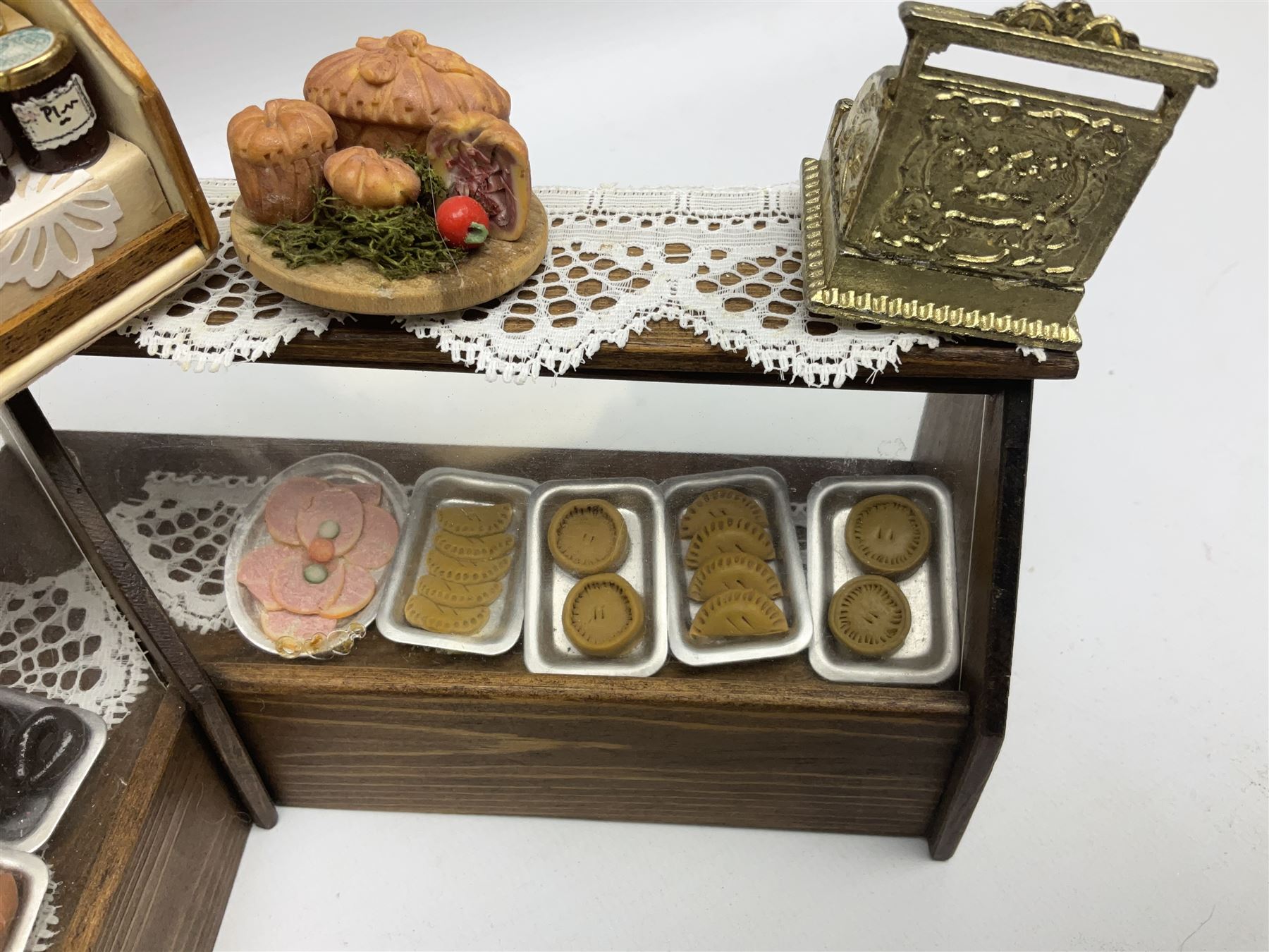 Collection of miniature dolls house grocery shop furniture and produce, comprising glazed corner counter with brass style cash register, shelved preserves unit, pies, cheese board and trays of meats, pair of white cheese counters, biscuit display with six tins, glazed preserves cabinet, pair of shop chairs and four further cheese boards complete with grapes and label flags