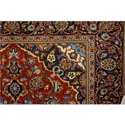 Central Persian Kashan hand-knotted rug, madder red field with central articulated cusped medallion in ivory and sky blue, issuing network of scrolling arabesques palmettes and floral sprays with conforming spandrels, indigo main border of scrolling vinery and stylized palmettes, framed by multiple guard borders, with cotton fringe ends  