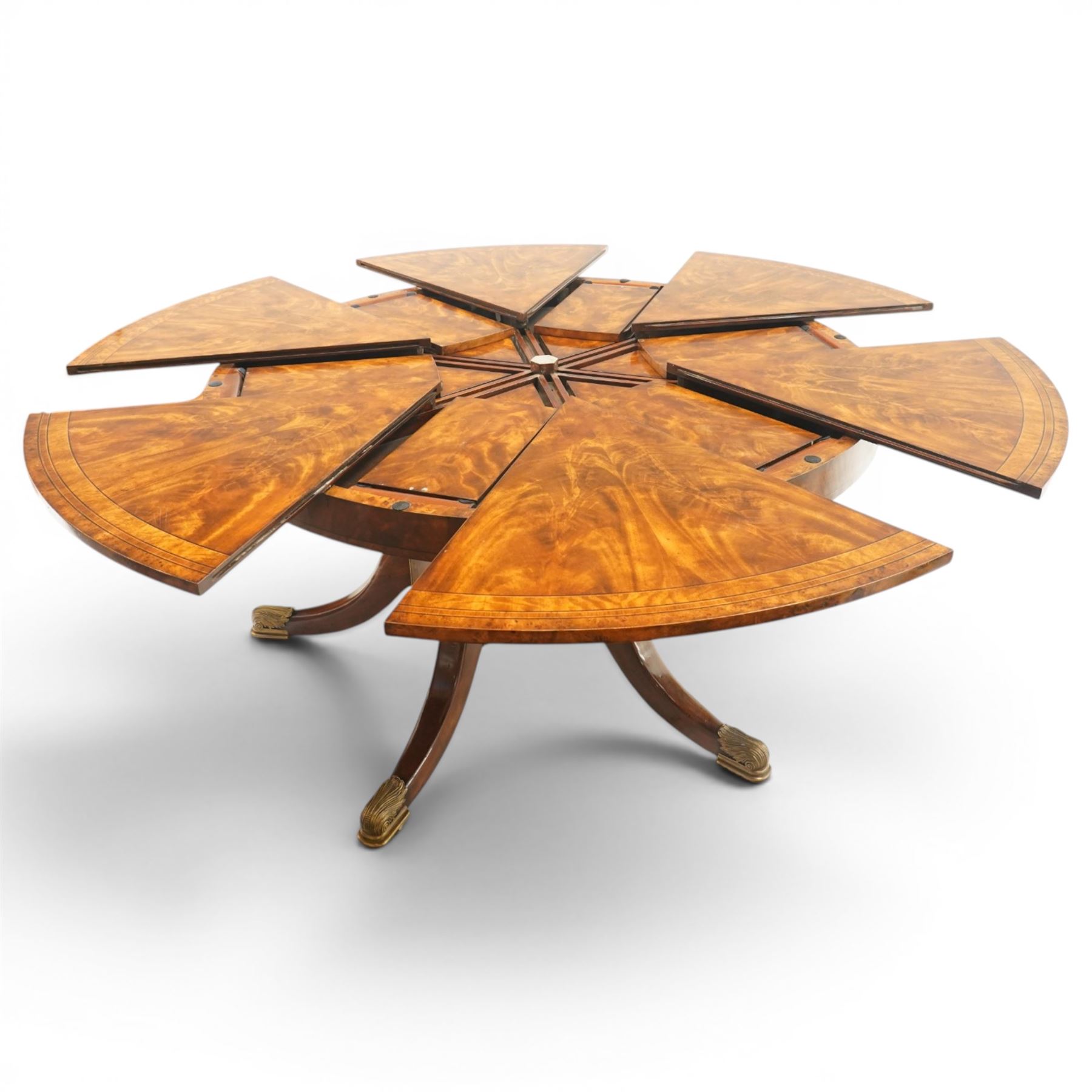 Theodore Alexander - Regency Revival figured mahogany 'Althorp Patent Jupe' extending dining table, the segmented circular top formed of six pull-out triangular divisions with inset fold-out leaves, raised on a large turned vasiform pedestal on a pentagonal platform base with ebonised stringing, terminating to five splayed supports with gilt metal acanthus scrolled feet, retailed by David Hick Interiors, Jersey