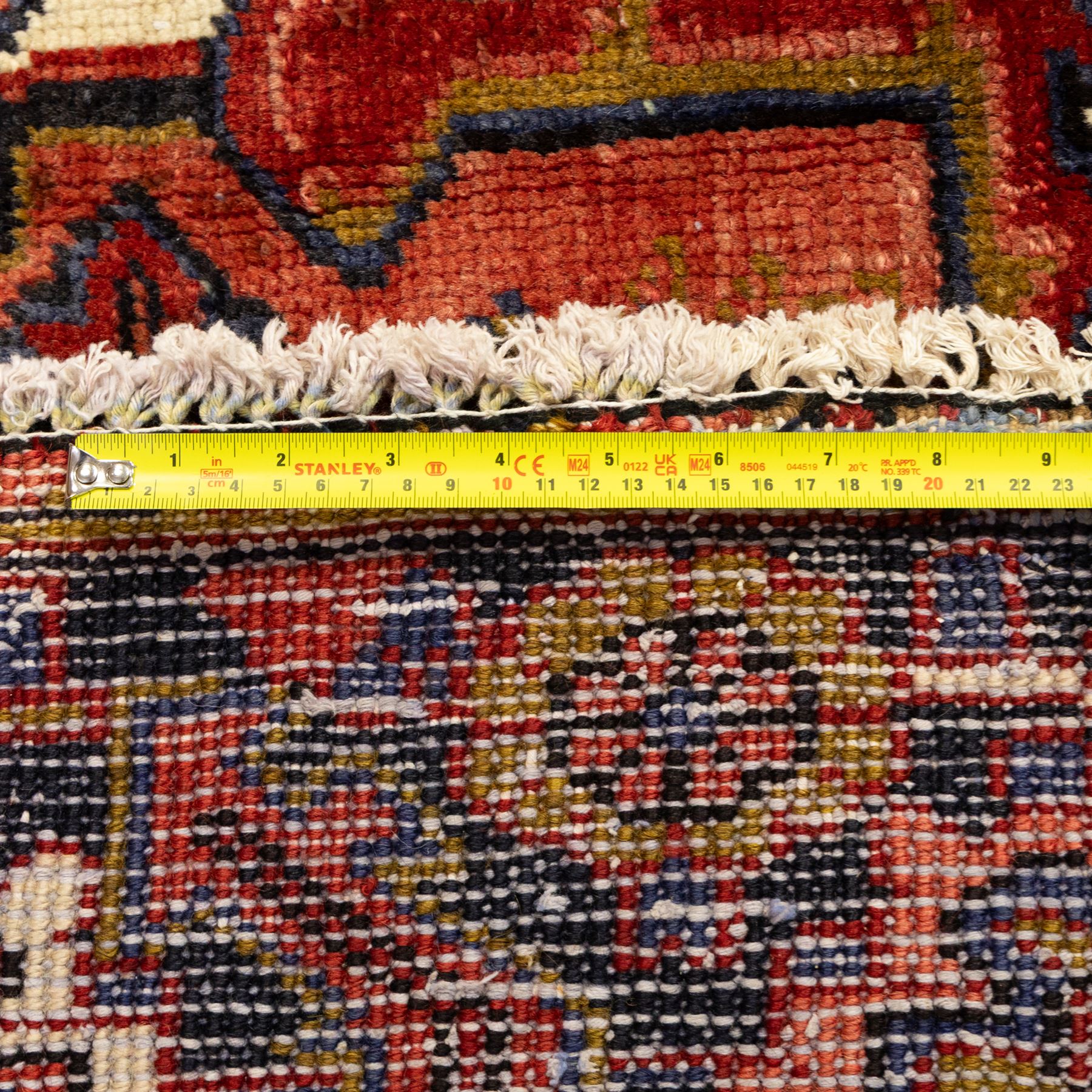 North West Persian Heriz hand-knotted carpet, madder red ground, with a large central geometric medallion with pendants, surrounded by angular foliate and floral motifs incorporating stylised rosettes, palmettes and serrated vine work, enclosed by ivory spandrels and a dark navy border with repeating geometric flowerheads and angular meandering vines, flanked by narrow patterned guard stripes, with cotton fringes