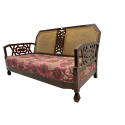 Early 20th century two seat sofa, cane back, the central panel and arm supports pierced with a geometric design, the sprung seat upholstered in fuchsia floral patterned fabric