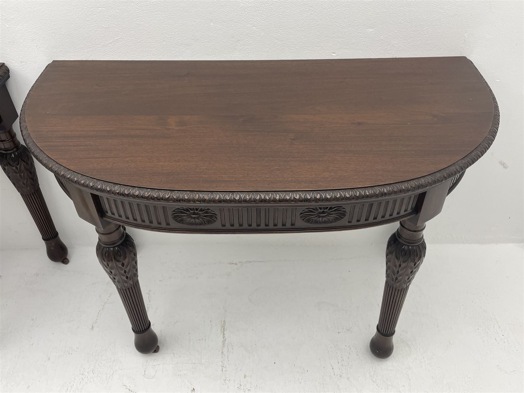 Pair early 20th century Hepplewhite style mahogany D-end console tables, the curved top with foliate carved edge, fluted frieze rails carved with flower heads, turned and reeded acanthus supports with recessed castors