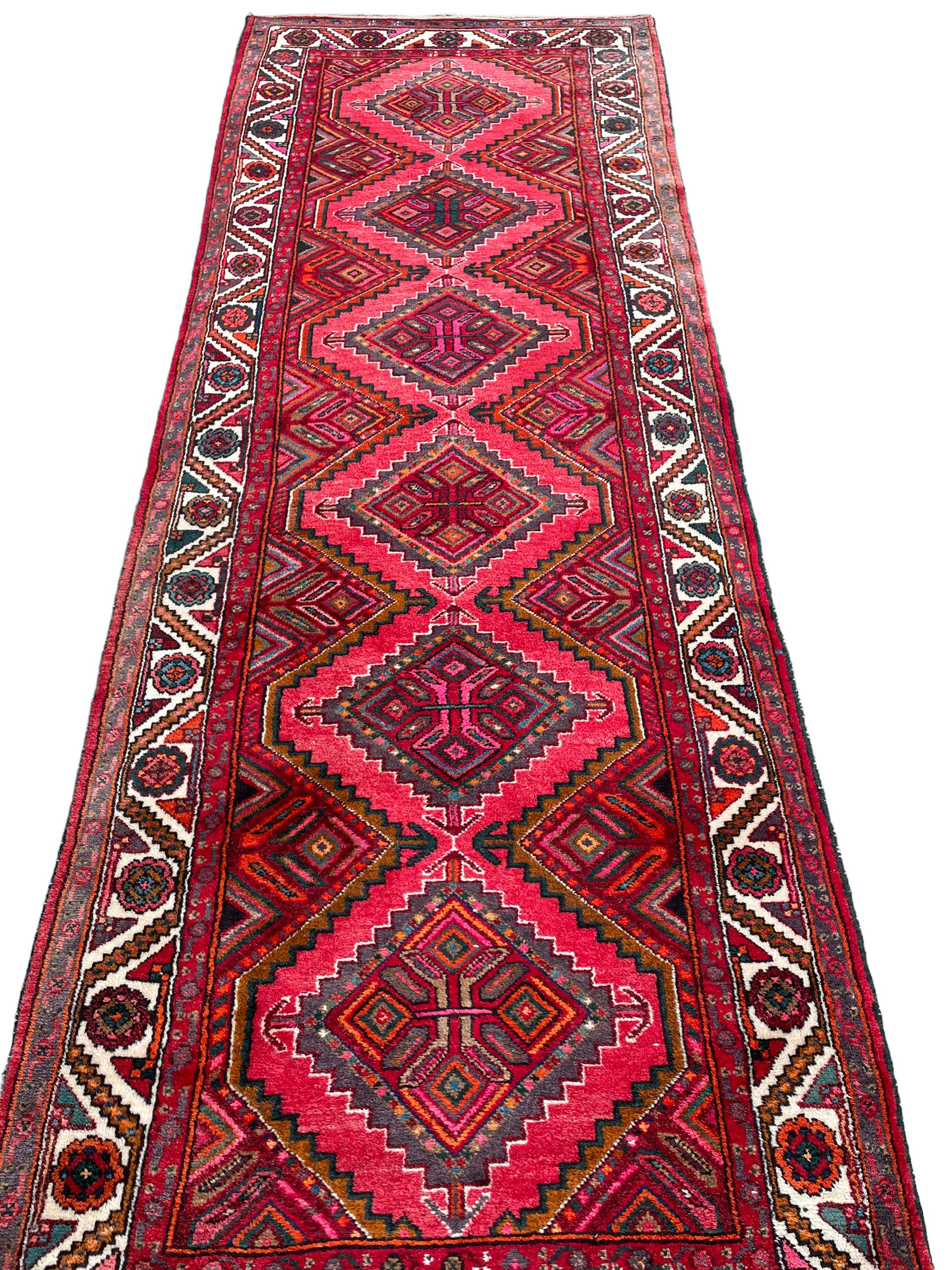 Persian Hamadan red ground rug, decorated with six stepped lozenge medallions, the guarded border with repeating stylised flower head decoration, with inscription to corner