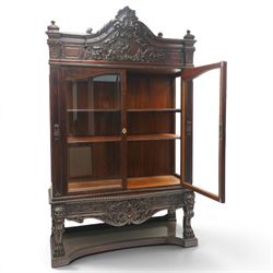Large and impressive 19th century heavily carved mahogany display cabinet, the ornate pediment carved with a central scalloped and foliate rocaille motif with extending acanthus leaves and egg-and-dart moulding flanked by acanthi finials, over a central cartouche with carved bow-and-arrow motif and trailing scrollwork, the bookcase enclosed by two glazed doors with bevelled glass sides, the base with a gadrooned upper edge over a central rocaille ornament with scrolling acanthus leaves, the panelled frieze with further foliate motifs, raised on large carved snarling lion mask supports with floral garlands and claw feet, united by a concave rectangular platform base with gadrooning egg-and-dart moulding