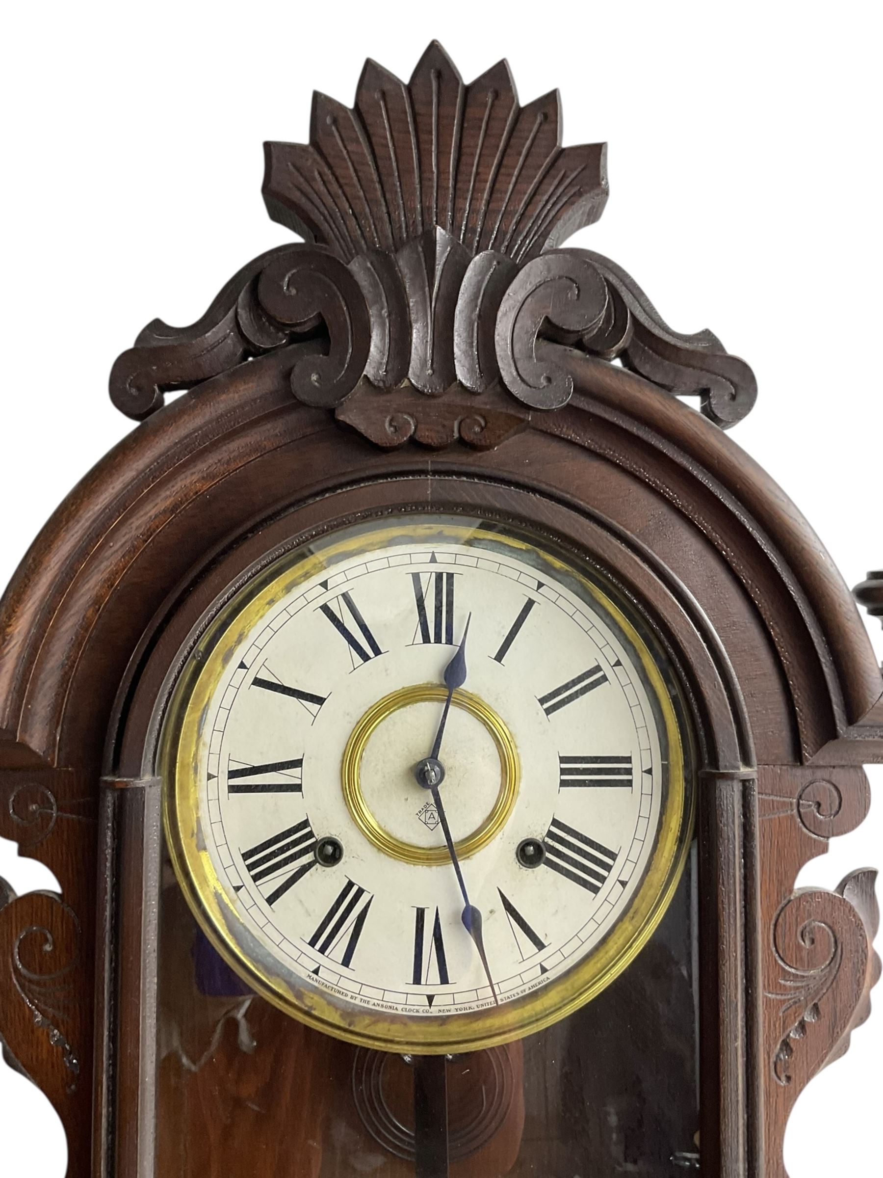 Ansonia-19th century American wall clock in a mahogany case with a carved pediment and base,  turned finials and carved side pieces, with a fully glazed door, dial with Roman numerals and visible pendulum, twin train spring driven movement striking the hours on a coiled gong. With key. 