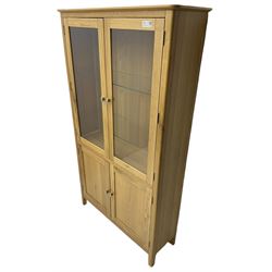 Contemporary light oak display cabinet on cupboard, fitted with two glazed doors enclosing two adjustable glass shelves with light fittings to the top, over panelled cupboard concealing single shelf, on square supports