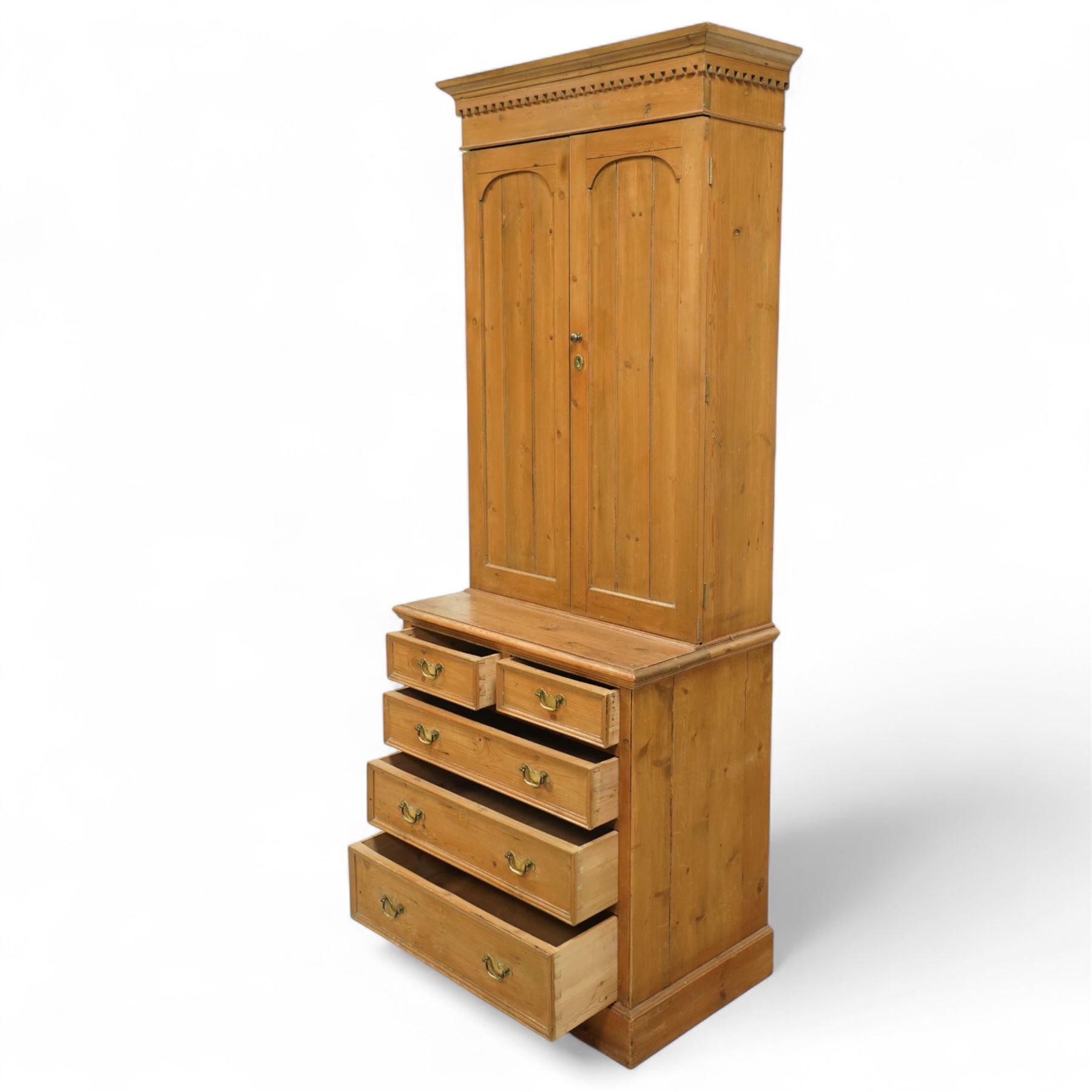Early 20th-century pine gun cabinet with later fitted shelves, of two-part construction, upper cupboard moulded and dentil cornice above two panelled doors revealing three removable shelves and circular recesses for gun barrels at the top, lower section with two short and three long graduated drawers with brass swan neck handles on plinth base 