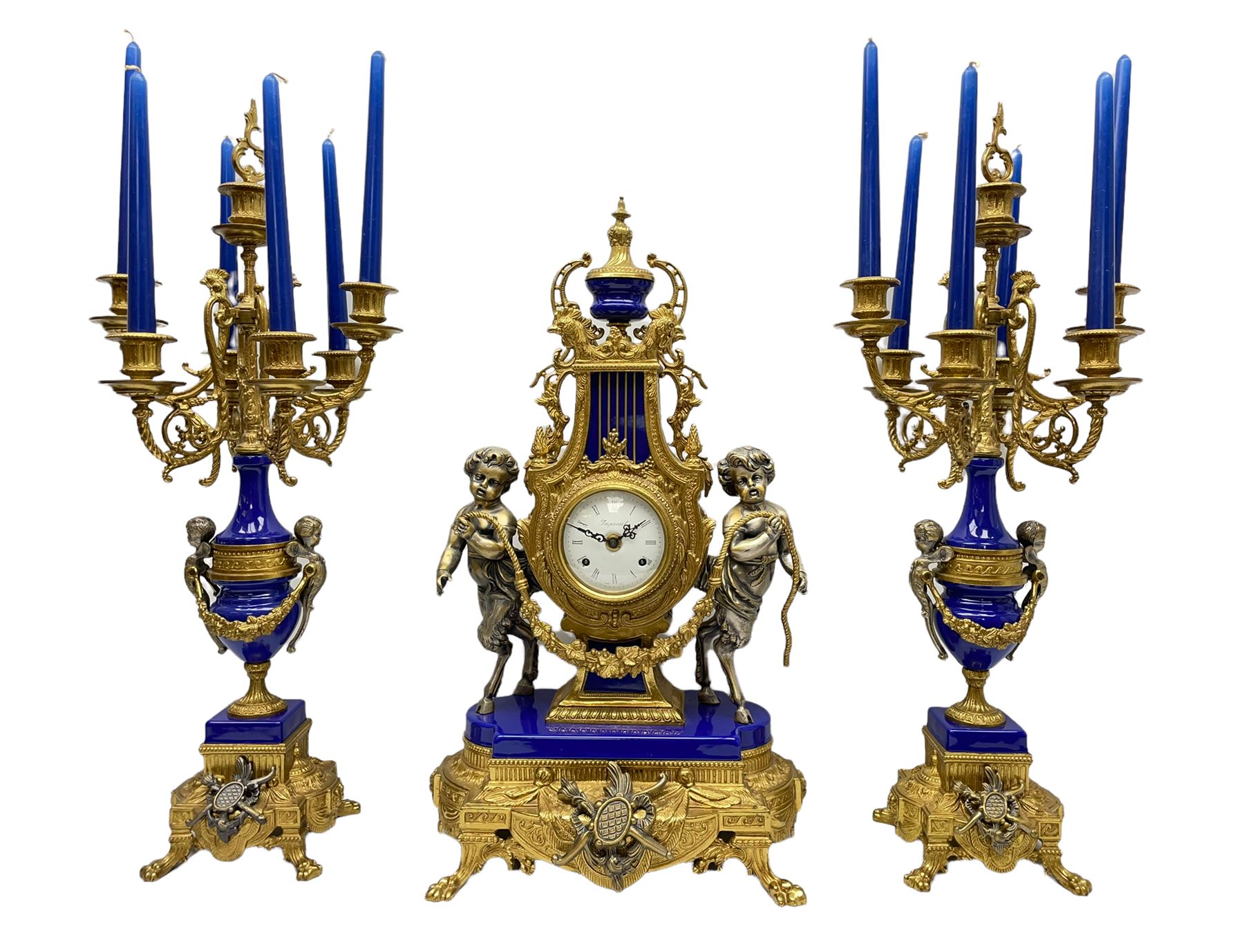 A 20th century continental gilt metal Lyre mantle clock with a pair of matching six light candelabra, spring driven movement housed in a gilt drum case on a brass and cobalt blue base with paw feet, two bronzed metal figures of mythological cherubs with animal legs and cloven hooves supporting a festooned garland swag, eight-day twin barrel striking movement with a floating lever balance escapement, striking the hours and half-hours on two bells, white enamel dial with roman numerals, minute track and pierced steel hands, dial inscribed “Imperial”. 