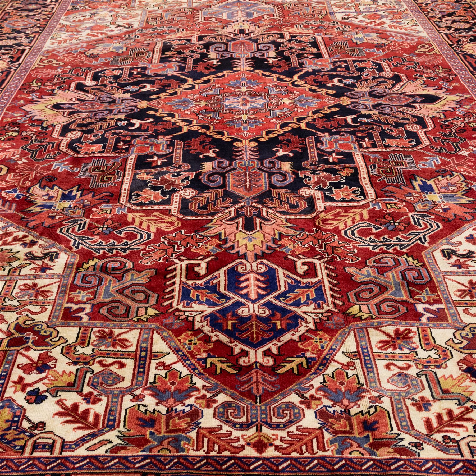 North West Persian Heriz hand-knotted carpet, madder red field with large central stepped medallion with angular floral and geometric designs, surrounded by stylized palmettes, leaves and tribal motifs with indigo corner spandrels, enclosed by a broad navy border of geometric floral devices between multiple guard stripes, with cotton fringe ends 