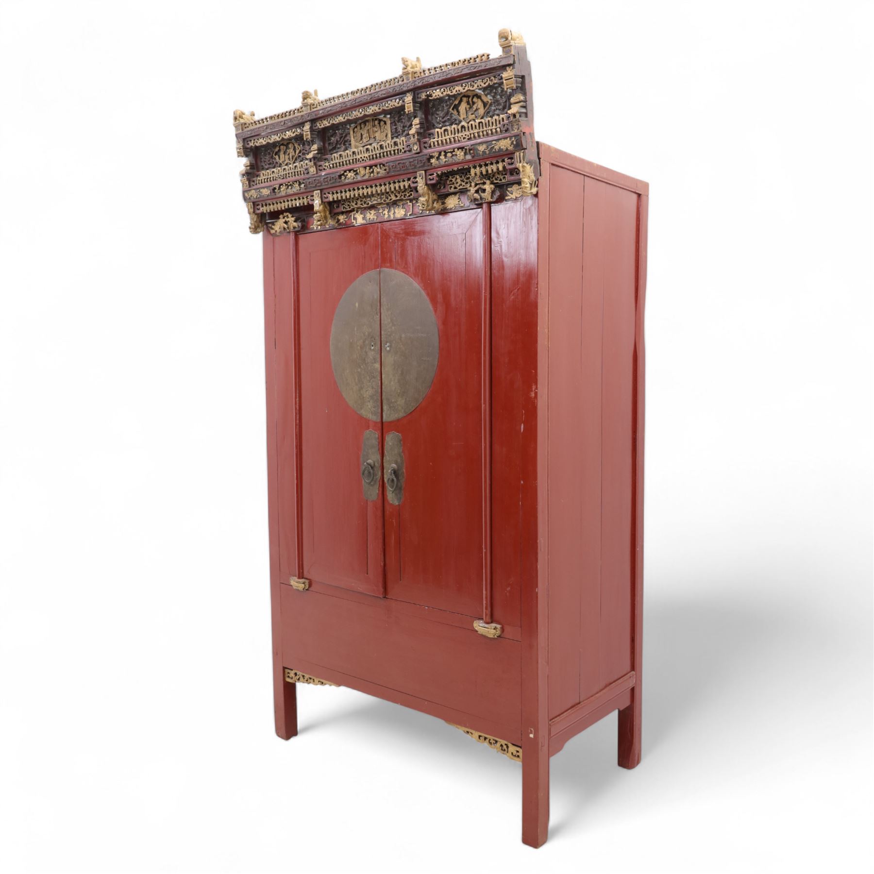Early 20th century Chinese lacquered marriage cabinet, of rectangular form in red lacquer, fitted with double doors centred by circular brass lock plate and drop handles, the interior with shelves and two short drawers, cornice and apron with giltwood foliate and figural carving, together with an elaborately pierced and gilded wooden cresting panel carved with scenes of figures within pavilions and stylised dragons