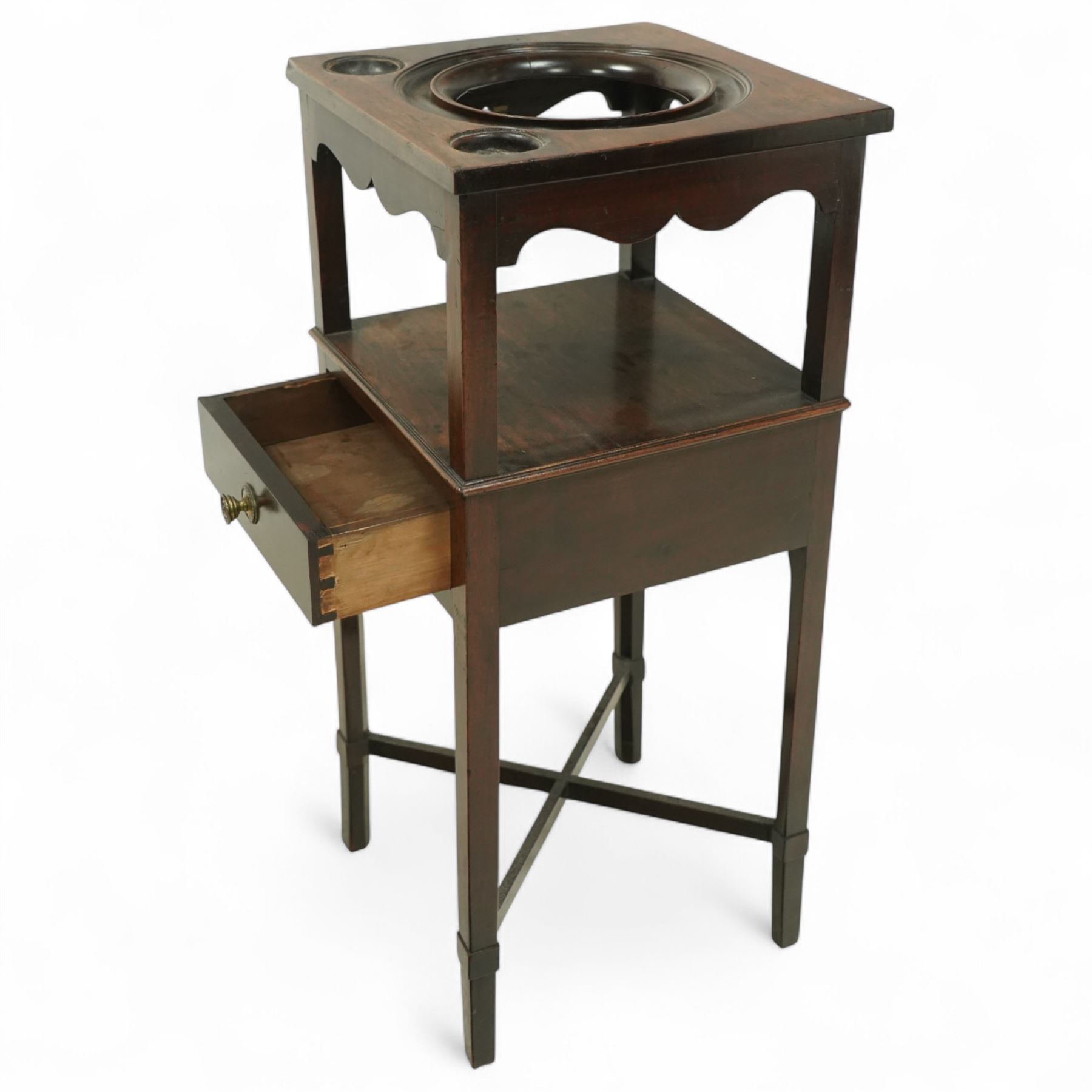 George III mahogany washstand, square top with circular wash bowl aperture and twin bottle recesses above a shaped frieze and single drawer with brass knob handle, raised on square tapered legs united by cross stretchers 
