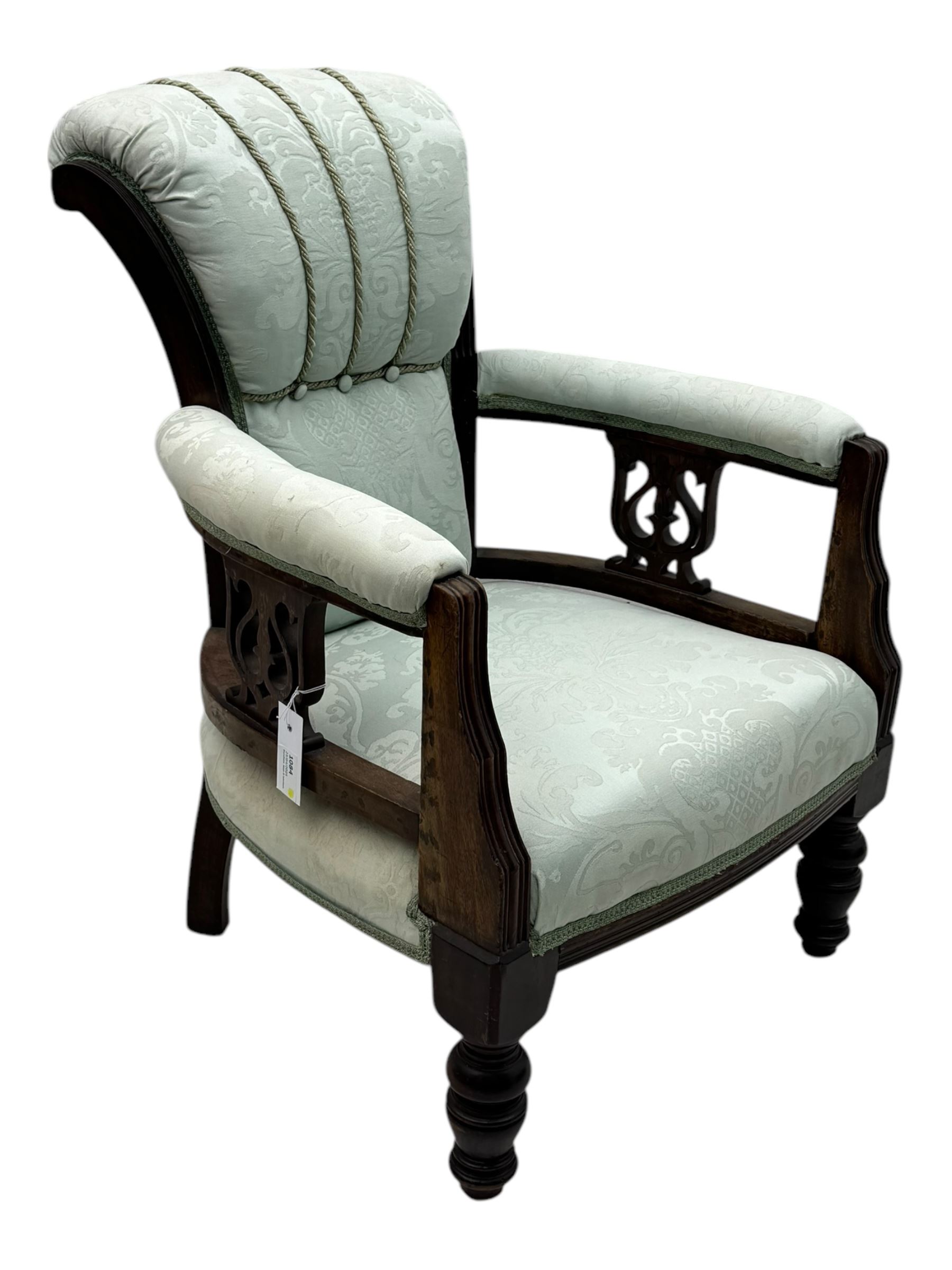 Late Victorian carved mahogany open armchair, the shaped back with fluted upholstered panel and padded arms above pierced scroll carved side supports, over a broad upholstered seat, on turned front supports