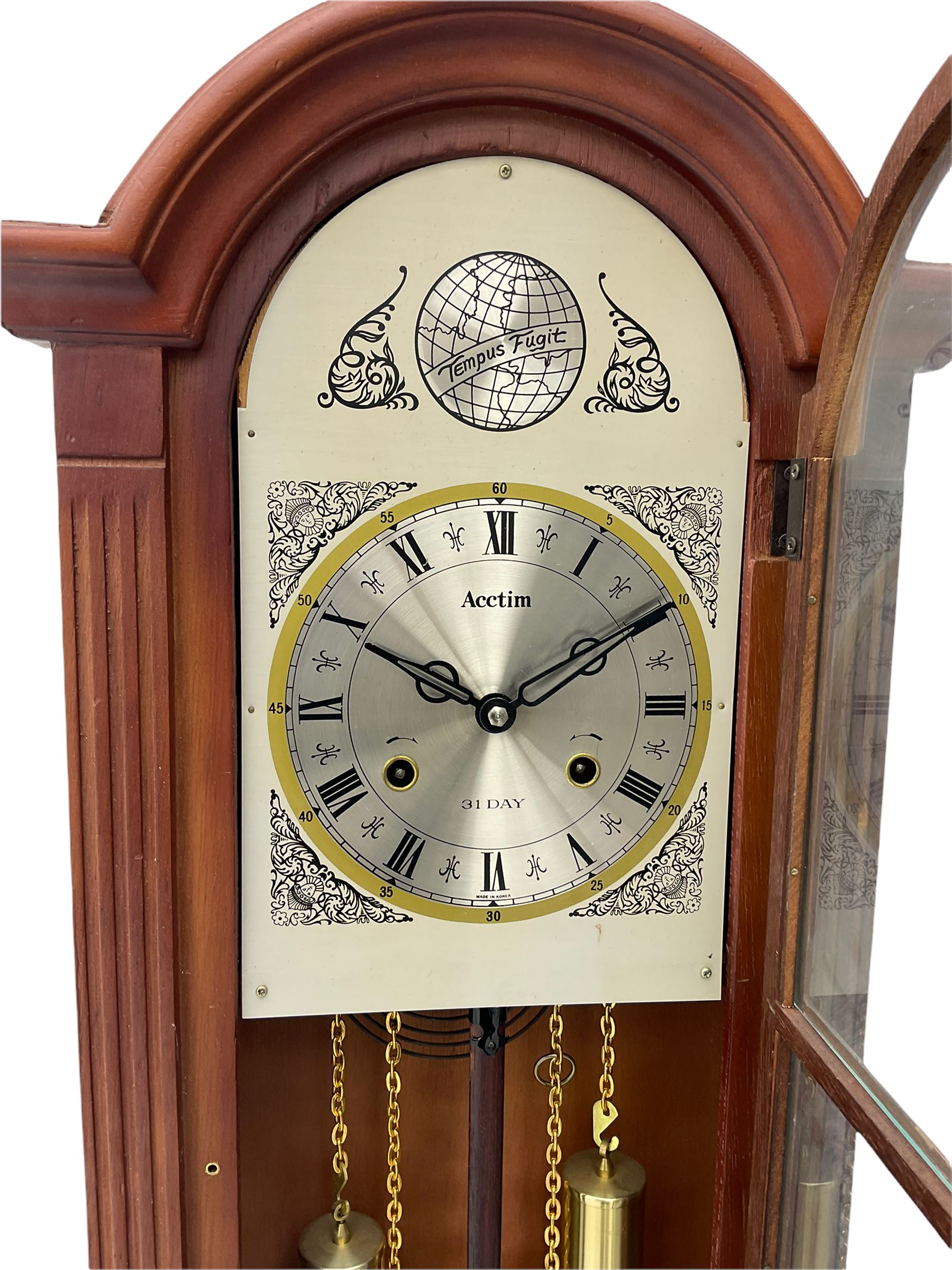 A contemporary Acctim (Anglo continental clock company) thirty-one day striking wall clock in a mahogany effect case with a fully glazed door displaying the pendulum and two dummy wights with chains, with a polished brass dial, Roman numerals, minute track and pierced steel hands, dial plate decorated with spandrels and Tempus Fugit inscribed on the dial brake-arch.
With key and pendulum.

