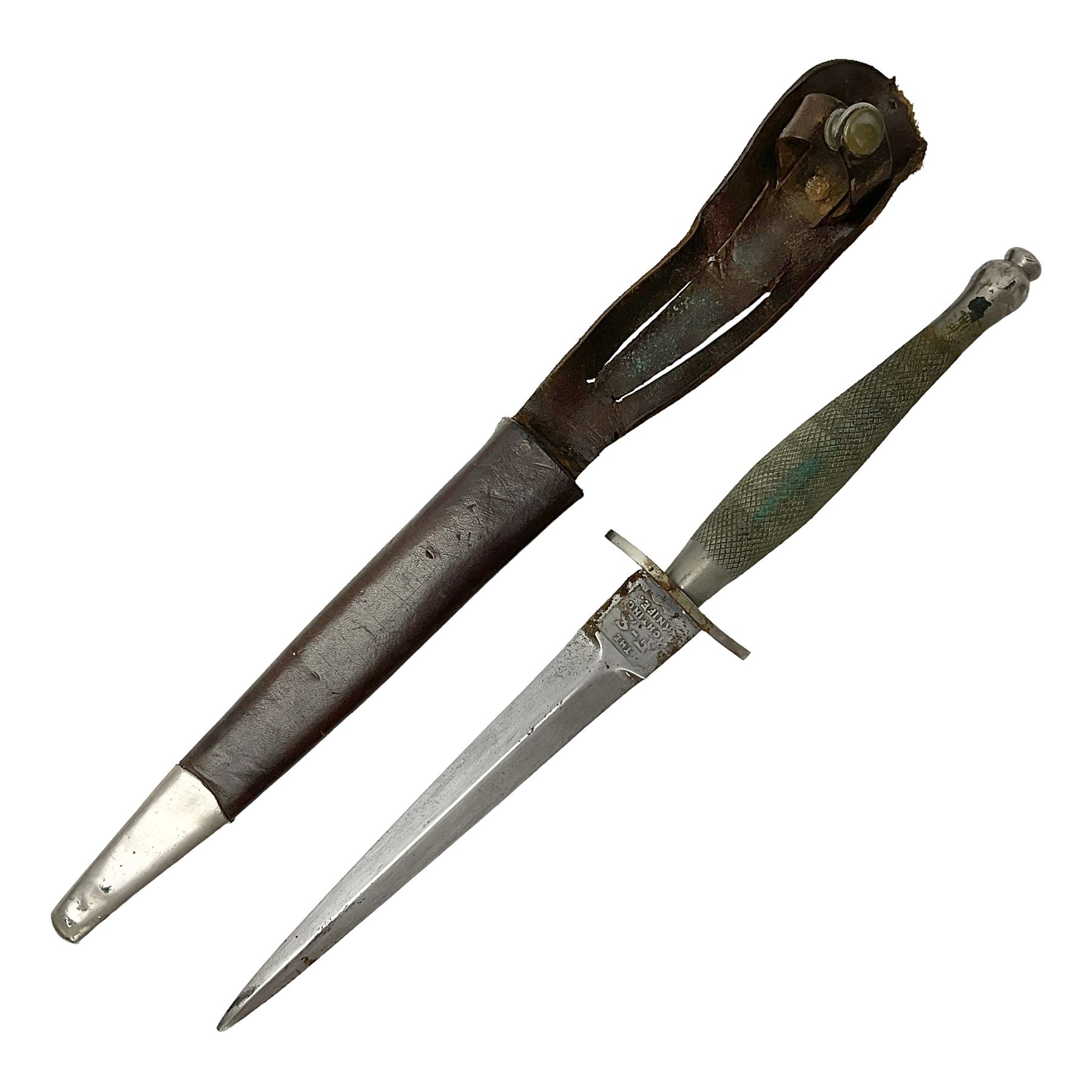 First Pattern Fairburn Sykes ''F-S'' Fighting Knife by Wilkinson Sword Ltd., the 17cm double edge steel blade with raised medial ridge to each side, each side of the ricasso with a recessed panel, one side with maker's name, the opposing side etched THE F~S FIGHTING KNIFE, with nickel plated brass recurving crossguard and diamond cut grip; in leather scabbard with metal chape