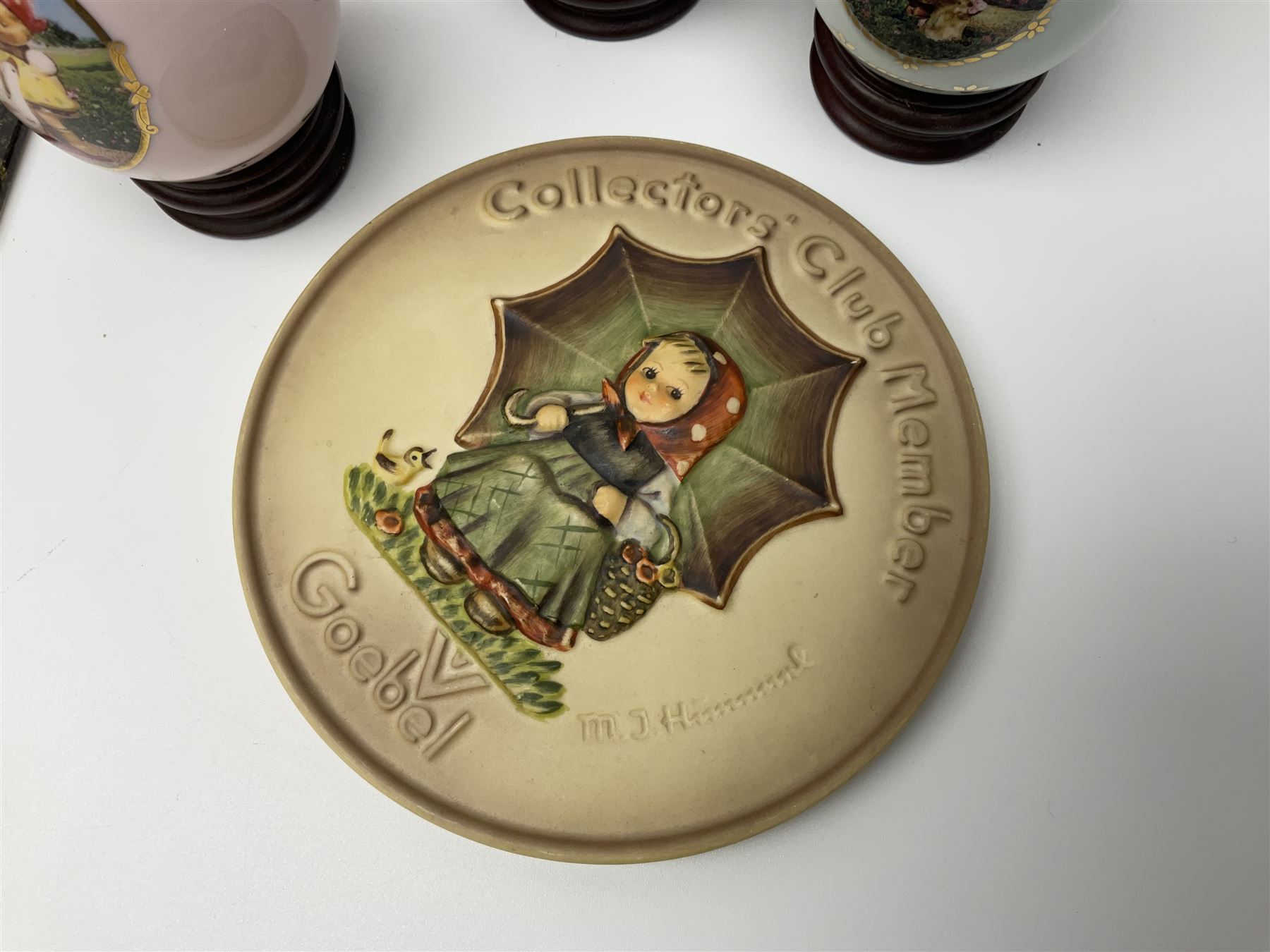 Two ceramic Hummel postcards by Goebel, both displayed on wooden easels, together nine Hummel eggs, set of four Hummel School Days plates, on wooden hanging rack, Goebel Collectors Club Member plaque etc