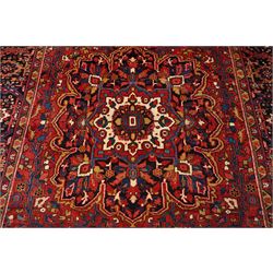 North West Persian Heriz/Ahar hand-knotted carpet, brick-red field with a large central angular stepped medallion with pendant motifs, surrounded by stylized palmettes, serrated leaves and geometric floral devices , ivory corner spandrels, enclosed by a dark indigo main border of stylized rosettes and angular vine motifs, between multiple guard stripes, with cotton fringe ends 