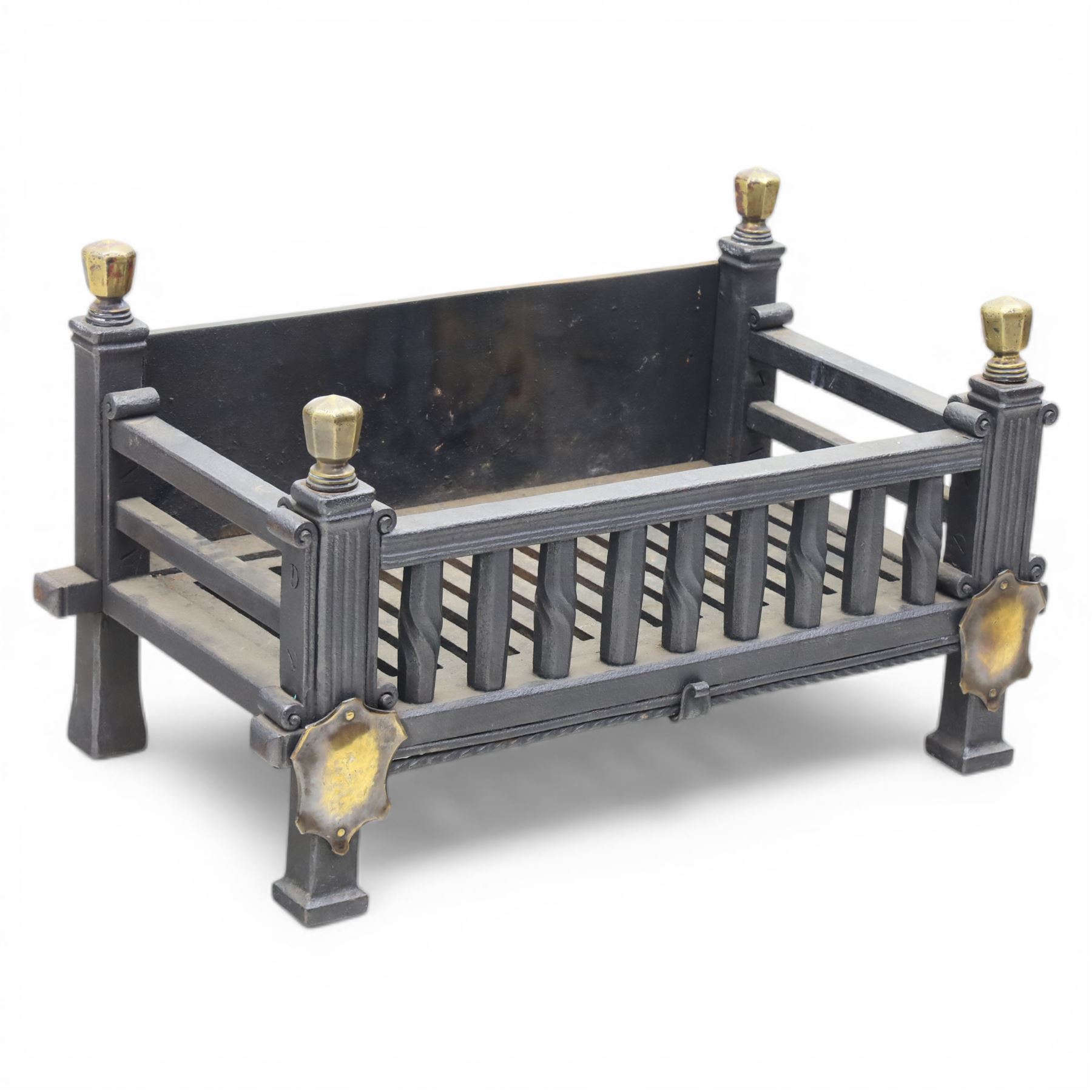 Arts and Crafts cast iron fire grate, rectangular backplate with plain upper rail over a slatted front of cylindrical tapering bars, flanked by square section uprights with fluted detail and scroll terminals, raised on stepped feet with shaped brass roundel mounts and brass finials to the corner posts