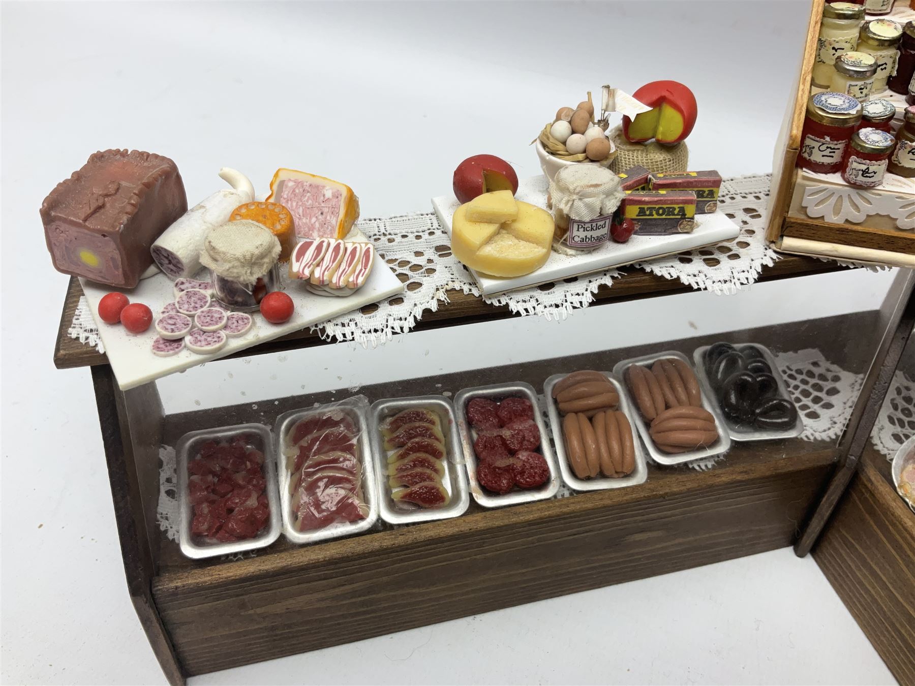Collection of miniature dolls house grocery shop furniture and produce, comprising glazed corner counter with brass style cash register, shelved preserves unit, pies, cheese board and trays of meats, pair of white cheese counters, biscuit display with six tins, glazed preserves cabinet, pair of shop chairs and four further cheese boards complete with grapes and label flags