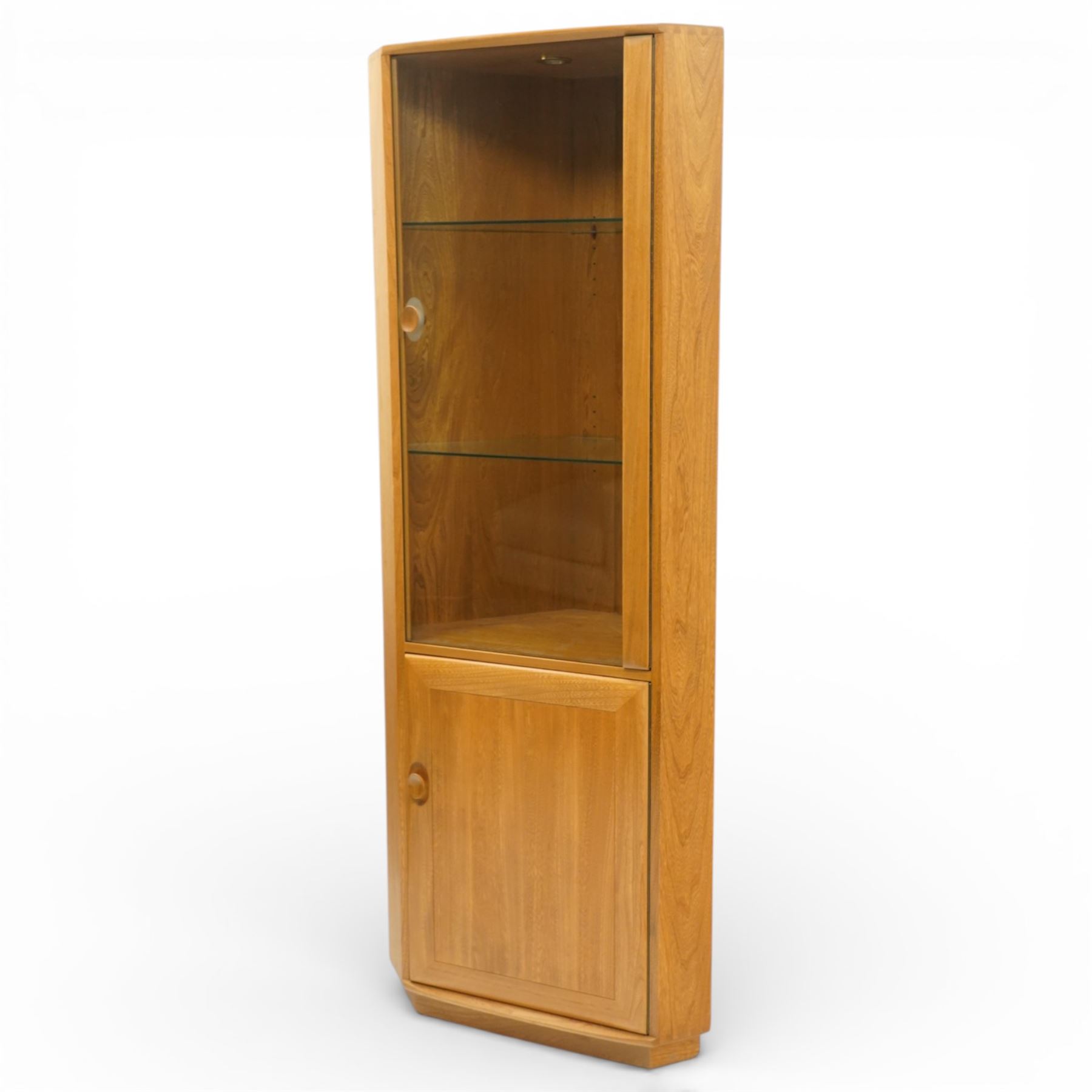 Ercol - elm corner display cabinet, Model 3856. Glazed upper section with glass door enclosing two glass shelves and interior light, above panel cupboard door, with rounded uprights and circular turned handles, on plinth base