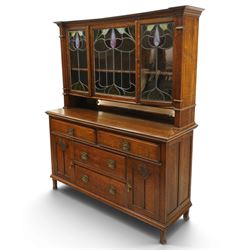 Arts and Crafts oak dresser, projecting moulded cornice above three glazed panels with coloured leaded glass floral panels, flanked by two glazed doors enclosing two adjustable shelves, open mirrored recess below with mirrored back, the base fitted with two short drawers over two central drawers flanked by carved panelled cupboard doors, ornate brass handles, raised on tapering square legs