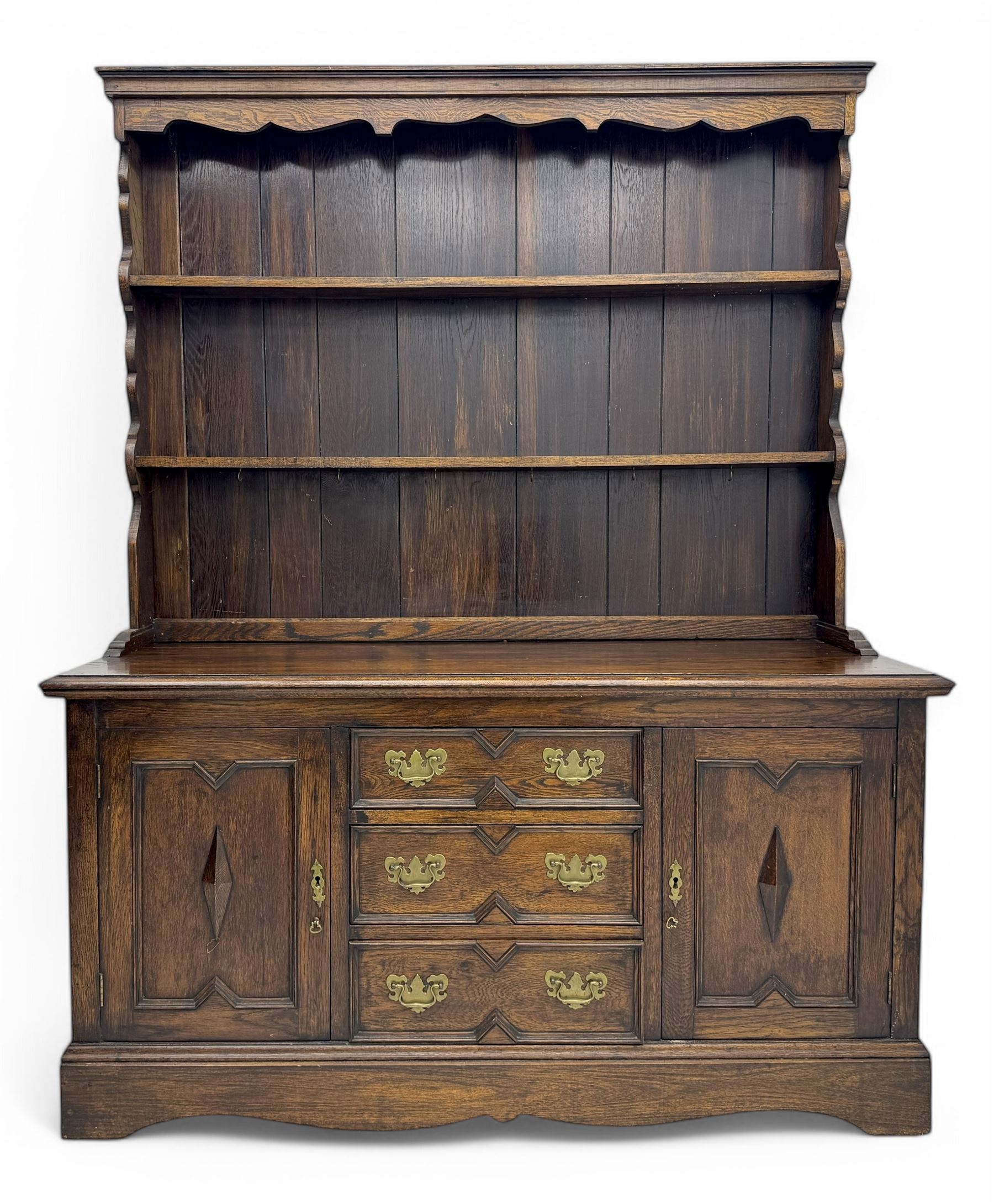 Late 19th century oak dresser, the upper section with two shelves beneath a shaped cornice, the lower section with a central bank of three drawers flanked by cupboard doors, all decorated with geometric diamond motifs, the drawers fitted with brass handles, raised on a shaped plinth base