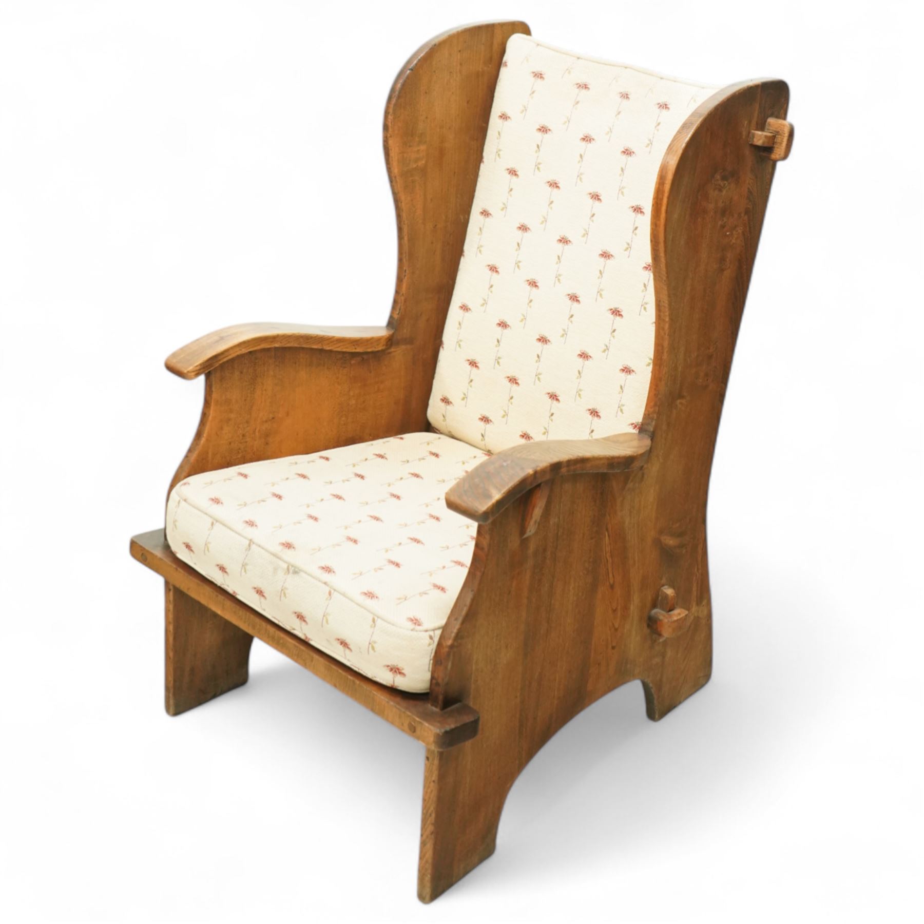 20th century pair of country lambing chairs in light oak, each with high winged back and shaped arms, pegged construction with panelled sides and arched aprons, loose seat and back cushions upholstered in cream floral fabric