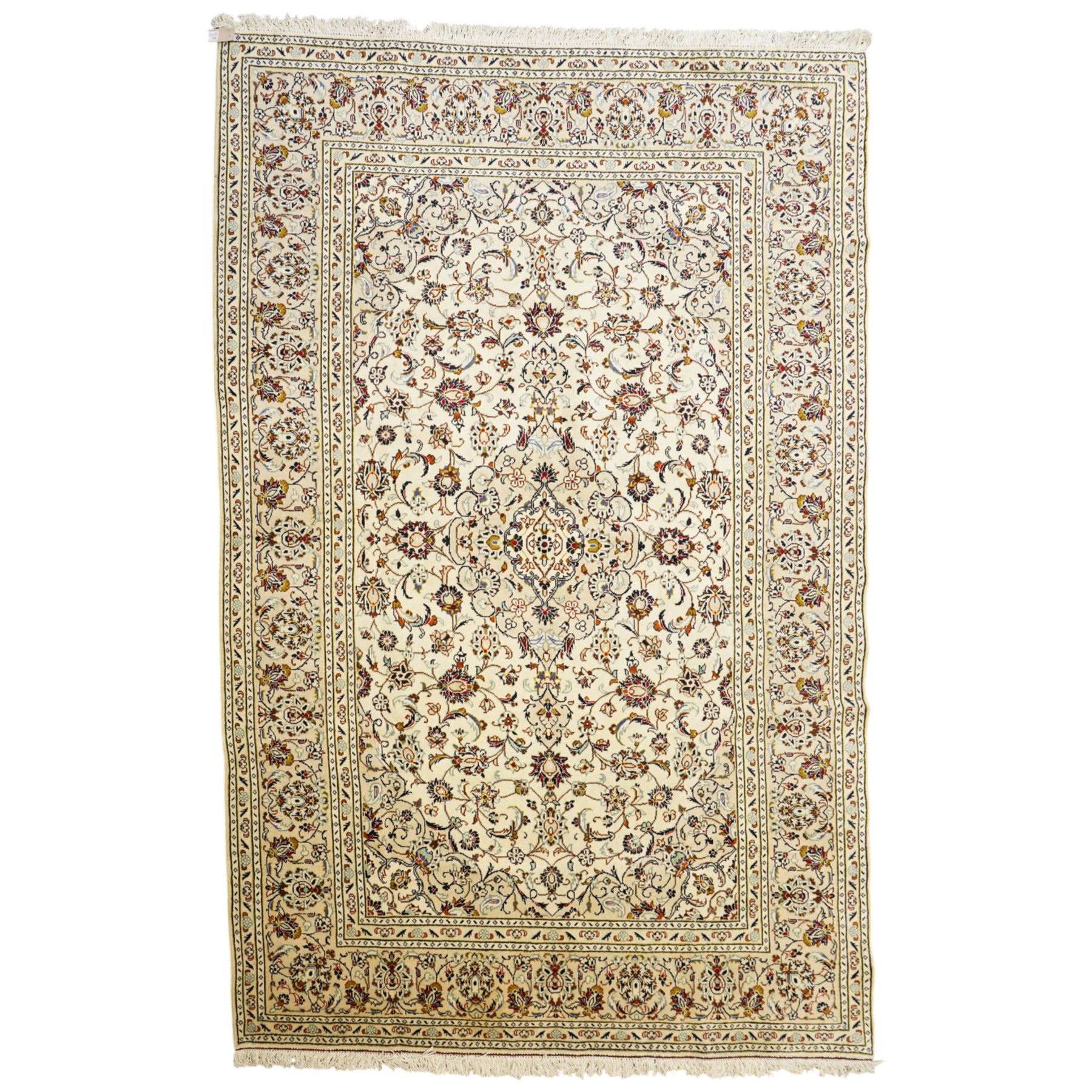 Central Persian Kashan hand-knotted carpet, ivory field with central lobed medallion issuing an all-over design of scrolling vines, palmettes, and floral sprays, within conforming spandrels, enclosed by a foliate border, framed by multiped guard stripes, with cotton fringe ends 