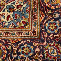 Central Persian Kashan hand-knotted carpet, crimson red central field with central floral medallion with floral vine-work and palmettes, within dense floral spandrels and a wide navy blue border decorated with continuous foliate motifs and multiple guard borders, with cotton fringe ends  