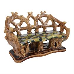 19th century pottery model of a bench, possibly Skey Pottery Tamworth, Staffordshire, the shaped seat mottled in shades of green, dark blue, yellow and brown, supported upon naturalistically modelled tree stumps, the arms and back modelled as intertwined branches, H14cm W21cm D10cm
