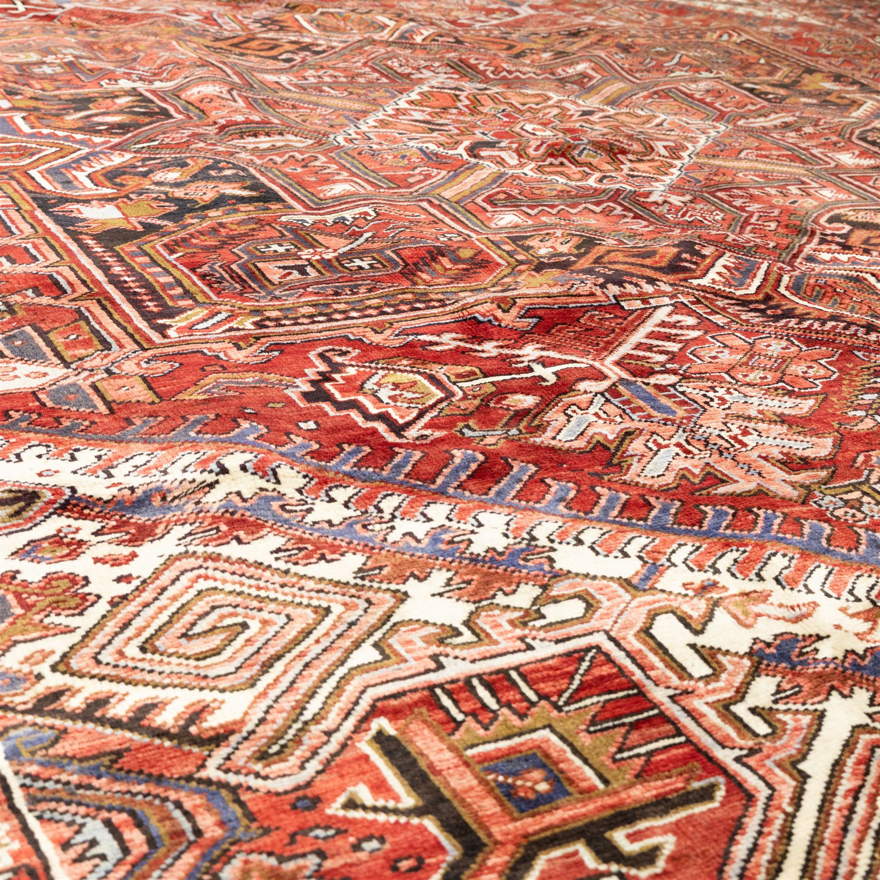 North West Persian Heriz hand-knotted carpet, madder red field, with a large angular stepped central medallion with serrated outlines, flanked by bold stylized palmettes, hooked leaves and geometric devices, spandrels similarly decorated within a wide geometric main border featuring angular floral motifs, enclosed by multiple guard borders in madder red, indigo blue, ivory, olive green and soft ochre, with cotton fringe ends 