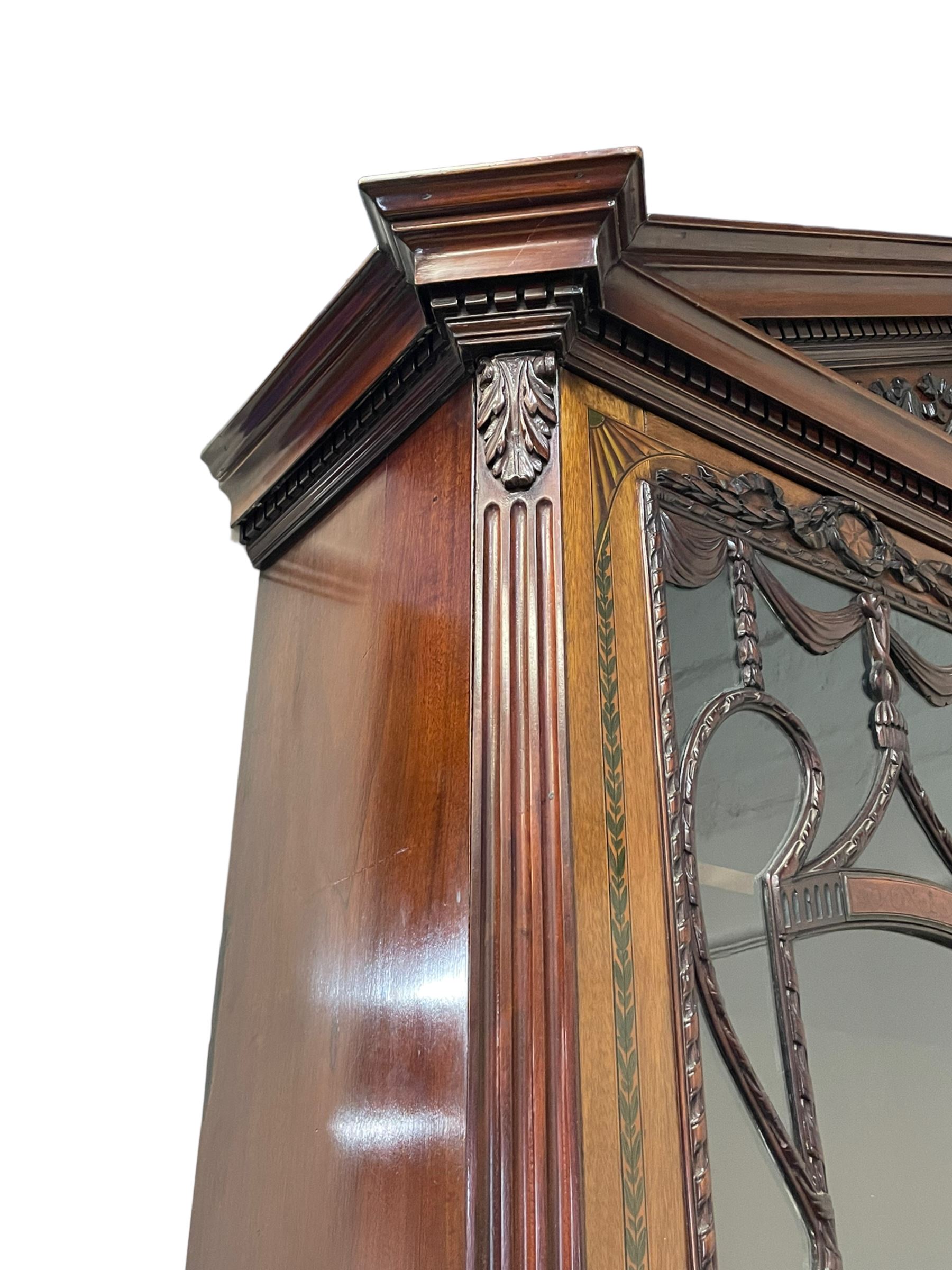 Large and impressive late 19th century Irish mahogany secretaire bookcase, moulded sloped arch dentil pediment decorated with central carved mask surrounded by linen festoons and trailing bellflowers, canted and fluted uprights with carved acanthus leaf upper and lower brackets, enclosed by two astragal glazed doors in the form of trophies with twist ribbon carved mouldings, inlaid with fanned spandrels and trailing leaf painted satinwood band, cylinder roll top inlaid with central fan motif and fanned spandrels, pull-out action revealing stepped fitted interior, central tambour roll cupboard flanked by two Composite pilasters and a combination of drawers and pigeon holes, fitted with three break-front drawers, matched canted and fluted corners with acanthus leaf carvings, on square tapering and fluted spade feet