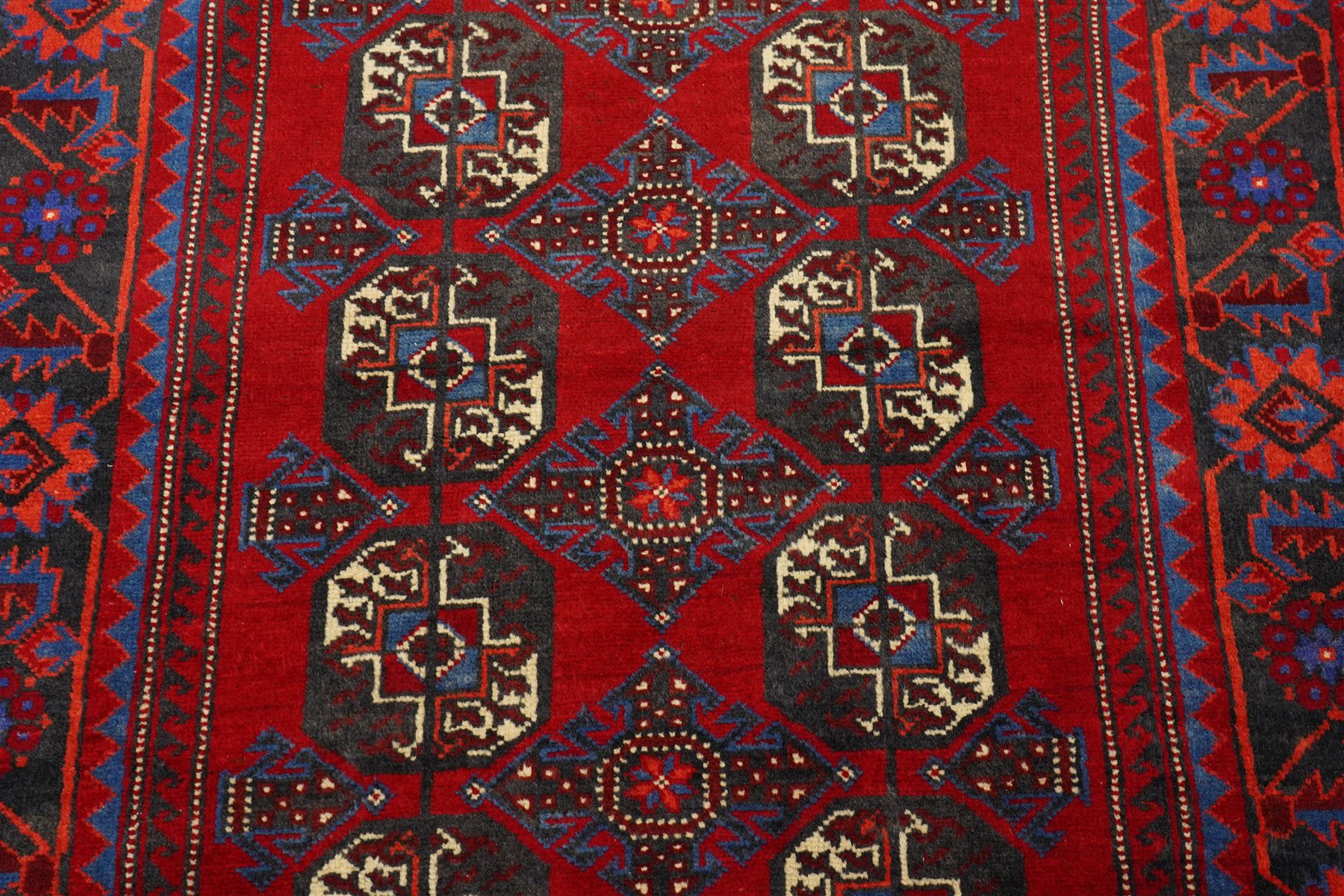 North East Persian Balochi hand-knotted runner, crimson field decorated with three vertical columns of repeating octagonal guls, each enclosing angular geometric motifs in deep indigo, ivory, and soft brown tones, guls linked by stepped diamond and cruciform devices, main border of stylized geometric floral motifs and angular vines, flanked by multiple guard borders, with cotton fringe ends 