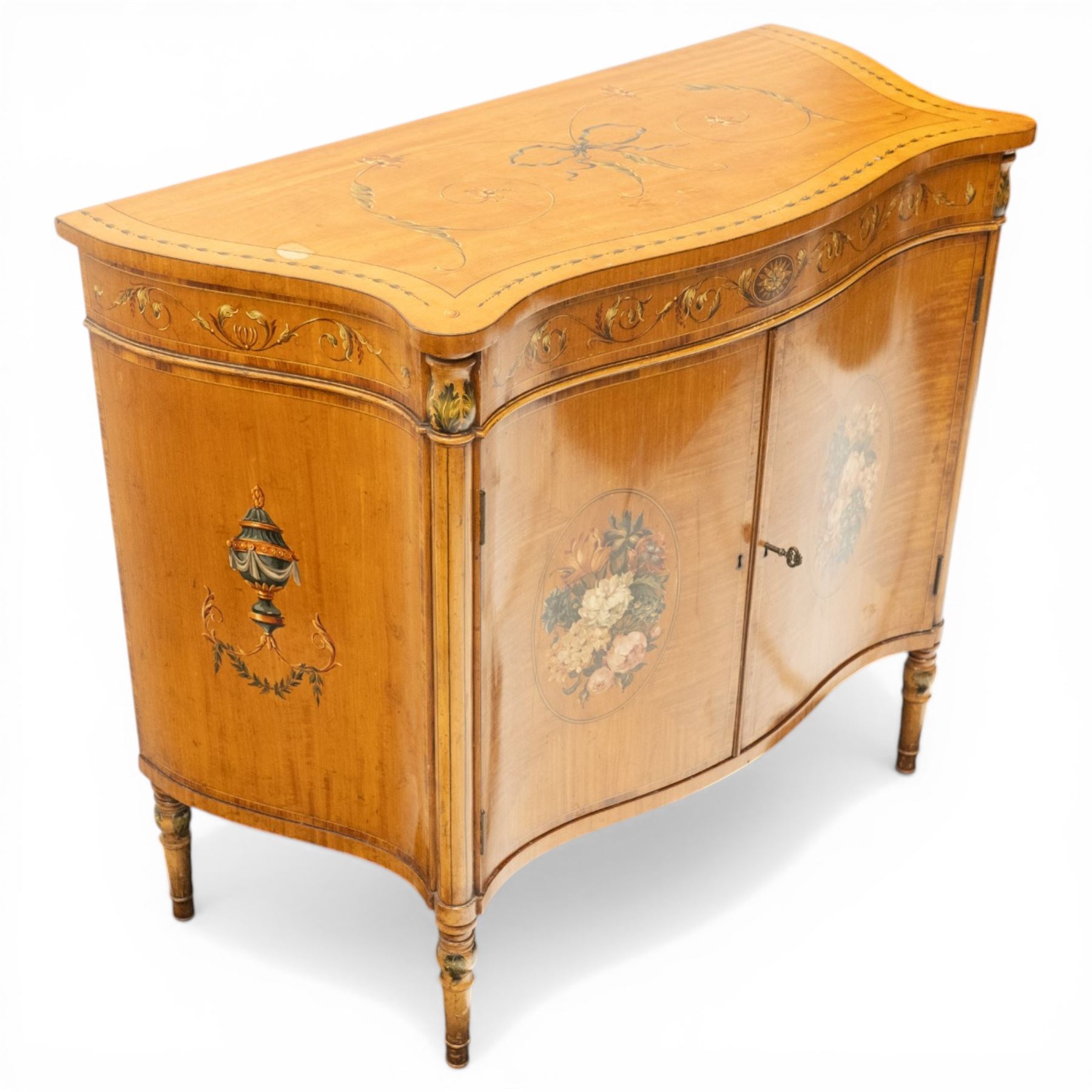 Sheraton Revival satinwood serpentine-front cabinet, the shaped cross-banded top with ebony stringing and painted scrolls of acanthi and ribbon work with a bell-flower border, the banded frieze painted with repeating acanthus scrolls, the matched veneer doors painted with oval panels of floral bouquets, raised on turned and tapering supports