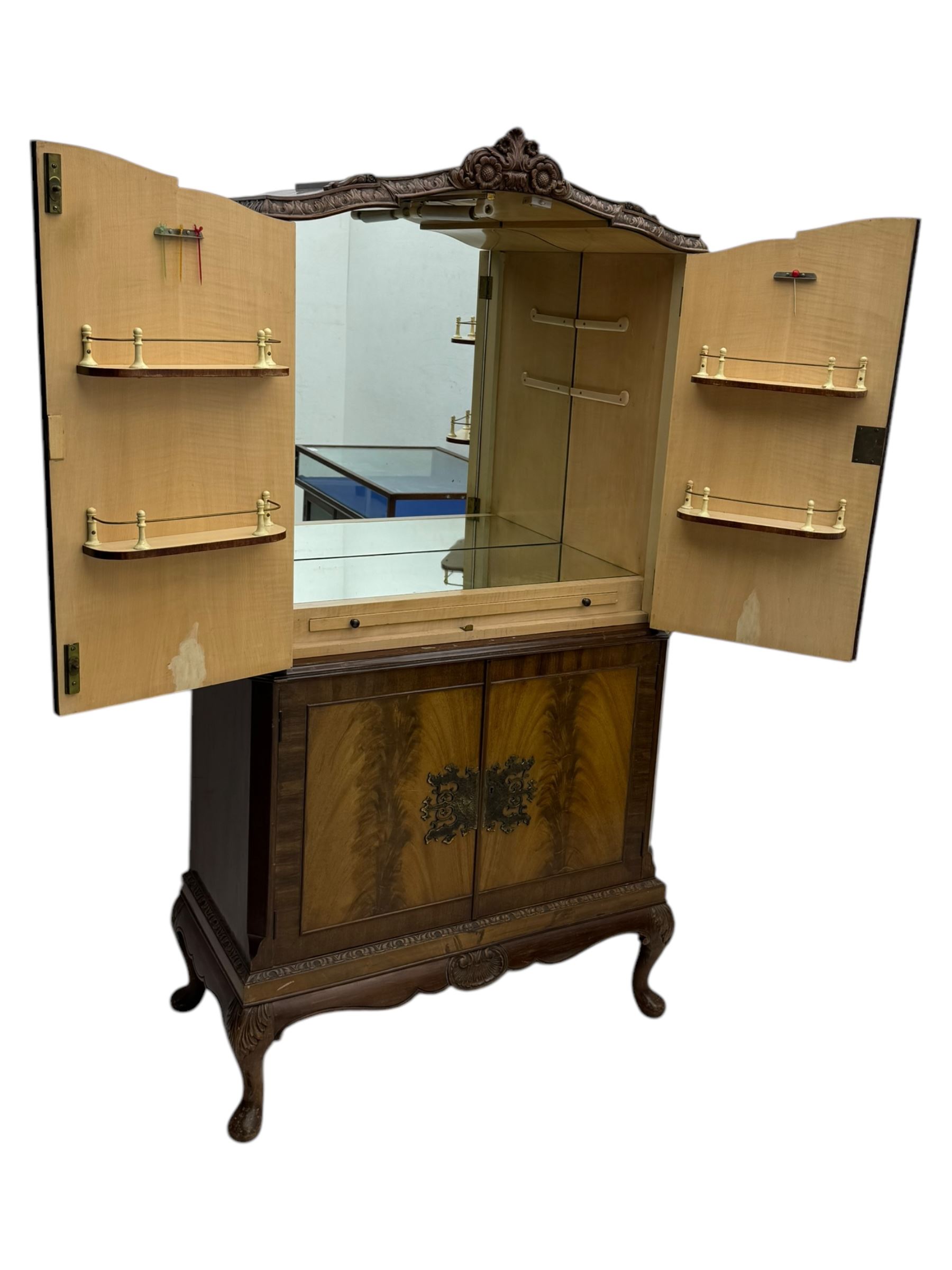 Early 20th century walnut veneered cocktail cabinet, shaped and carved foliate pediment above a pair of arched panelled doors with figured veneers and ornate cast escutcheons, interior is mirrored on the base and back, slide out mirrored shelf, flanked by foliate carved uprights, over a conforming lower cabinet, all raised on shaped apron and scroll carved cabriole feet