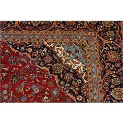 Central Persian Kashan hand-knotted carpet, crimson ground field, with central cusped lozenge medallion, enclosing a palmette and a floral rosette arrangement, surrounding field densely populated with scrolling arabesques, flowering vinery and stylized boteh motifs in tones of indigo, pale blue, ivory and ochre and spandrels of confirming design, flanked by multiple guard stripes, with cotton fringe ends 