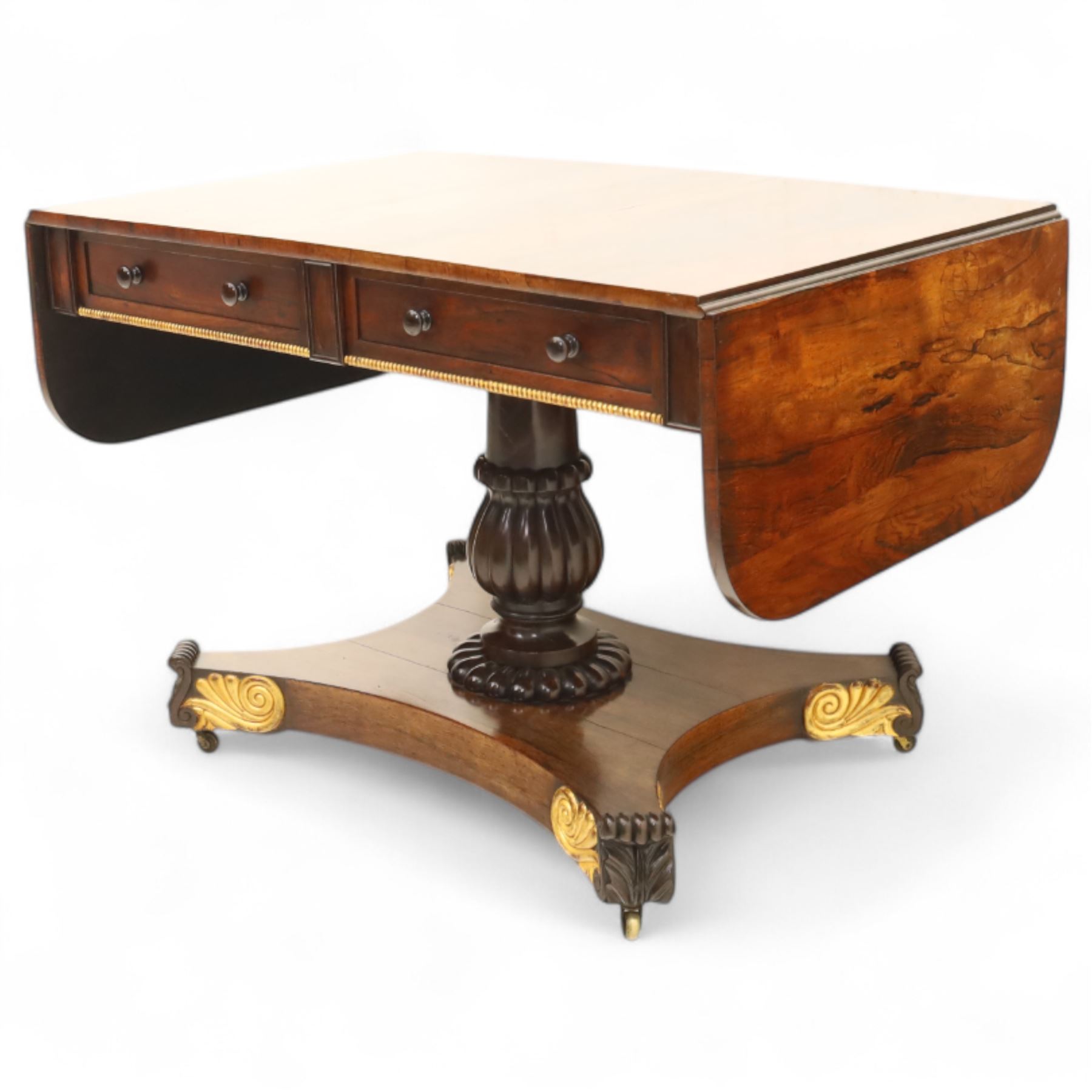 Regency rosewood sofa table, rectangular top with variegated rosewood veneers, hinged D-shaped leaves, over a set of dummy drawers to the back and full-size drawers to the front, with turned knob handles and gilt beaded moulding on frieze above elegant turned, tapering, and fluted, pedestal with a well-carved collar, supported on a large, tripartite concave platform base terminating in richly carved lion paw feet with gilded acanthus leaf carving, on concealed brass castors, circa 1815, length extended 161cm 

