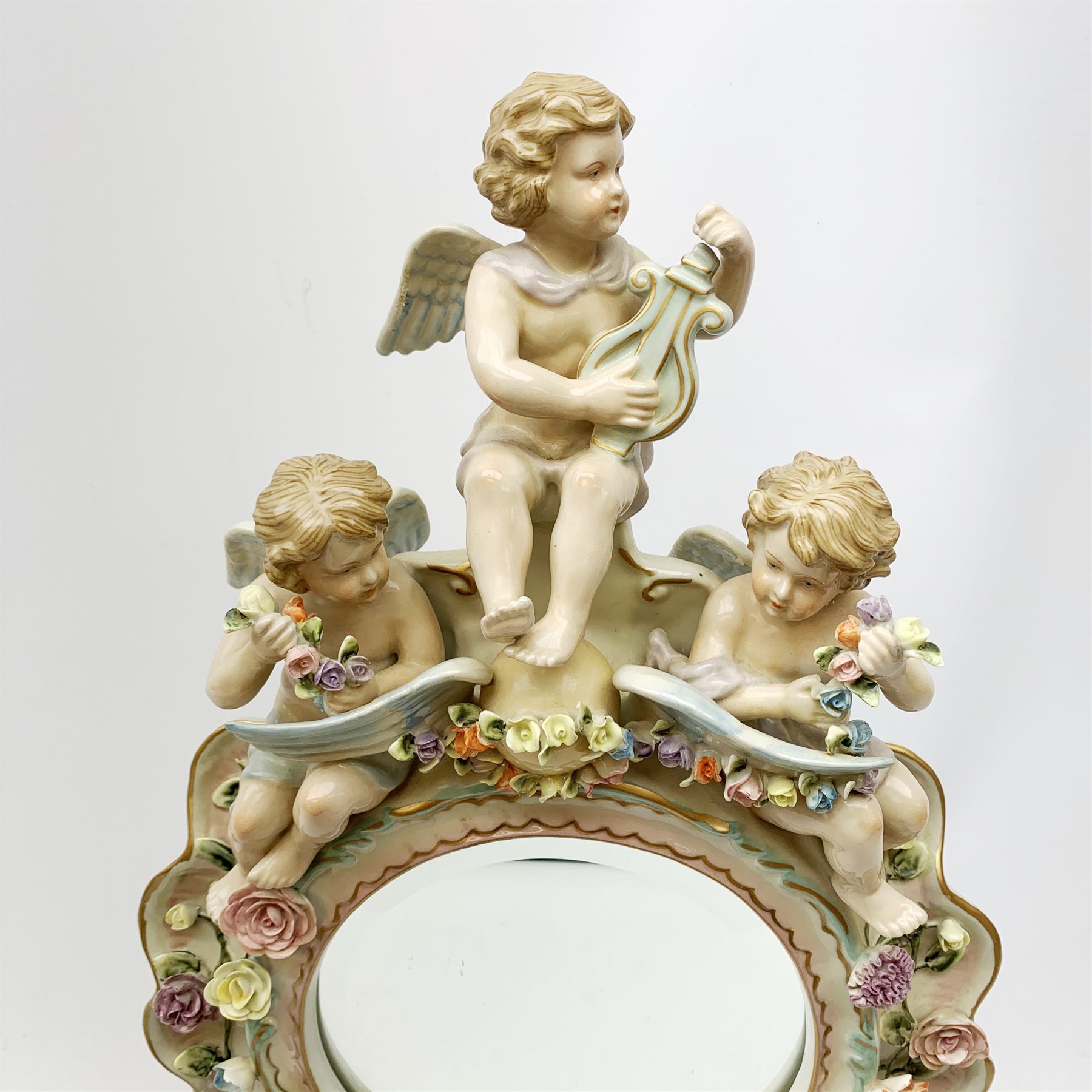  A late 19th century Sitzendorf porcelain mirror, the mirror plate of circular form set within a shaped surround encrusted with flowers and foliage, surmounted by three figures of cherubs and raised upon four scroll feet, with blue cross hatch mark beneath, H57cm.   