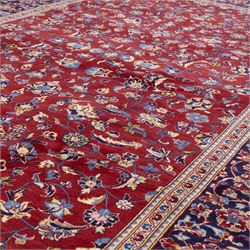 Central Persian Kashan hand-knotted rug, crimson field, decorated with an all-over floral and foliate design incorporating stylized palmettes, rosettes, and scrolling vine work, enclosed by a dark navy blue main border ornamented with flowering branches and palmettes flanked by multiple narrow guard borders with repeating floral motifs, with cotton fringe ends 