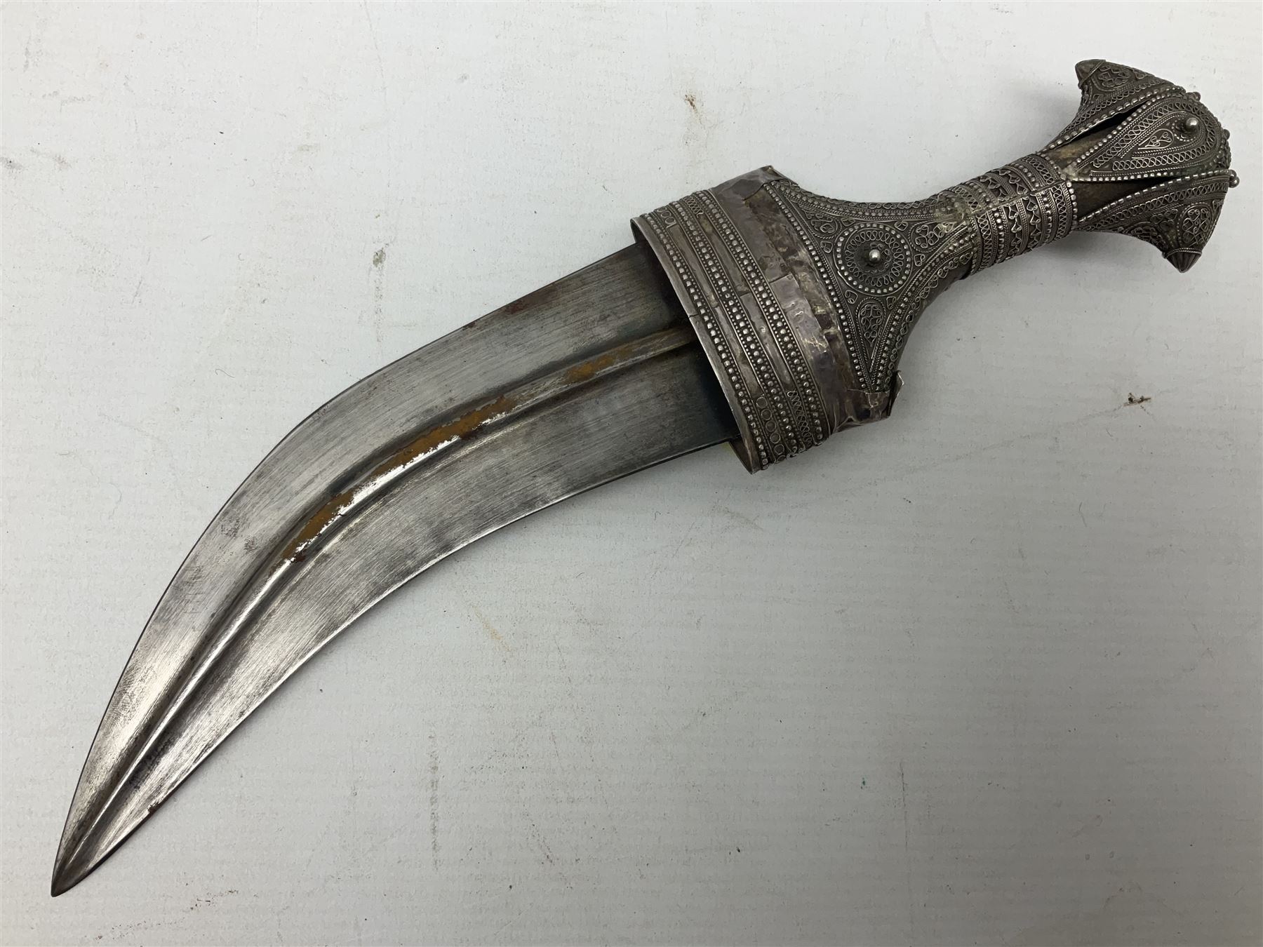 Omani Khanjar dagger, the 19cm curved blade with pronounced medial rib to either side, waisted hilt with dome shaped pommel and silver covered horn grip decorated with filigree bands, floral motifs and beadwork; in an intricately filigree decorated silver and leather scabbard with silver rings woven into the belt mount and fitted with original silver thread-work belt L34cm overall belt length 94cm