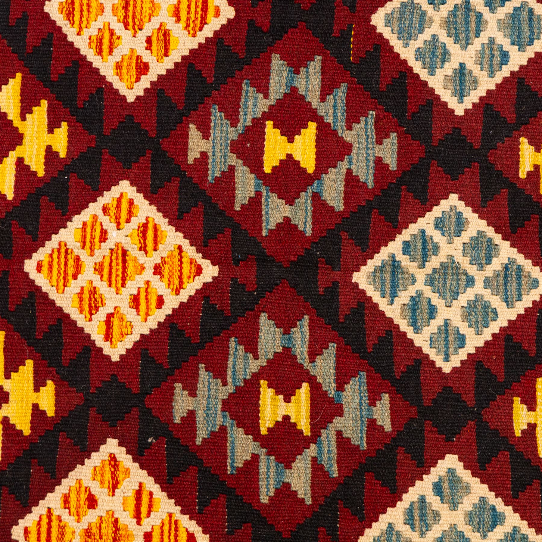 Afghan Maimana flatweave kilim, central crimson field features a large bold nested diamond motif and stylized geometric lozenge patterns on olive purple, blue, yellow and green tone ground, border stripe in teal ground, and fringe