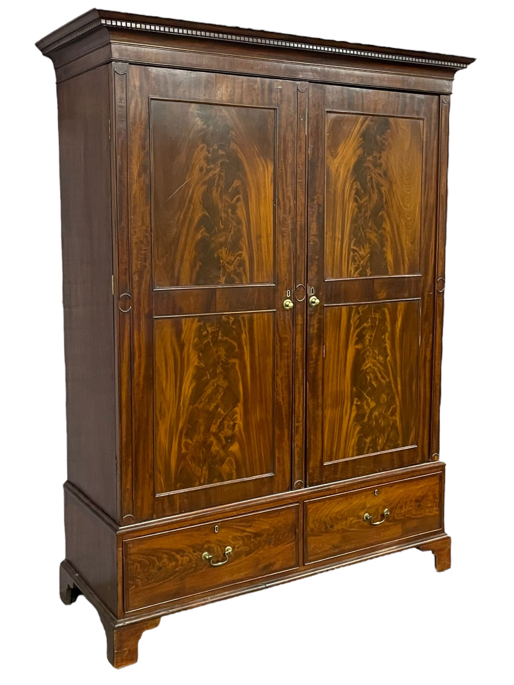 George III mahogany double wardrobe, the projecting cornice with dentil moulding over two panelled doors with mirror-lined interiors, enclosing hanging rails and brass hooks, the base fitted with two drawers, raised on bracket feet