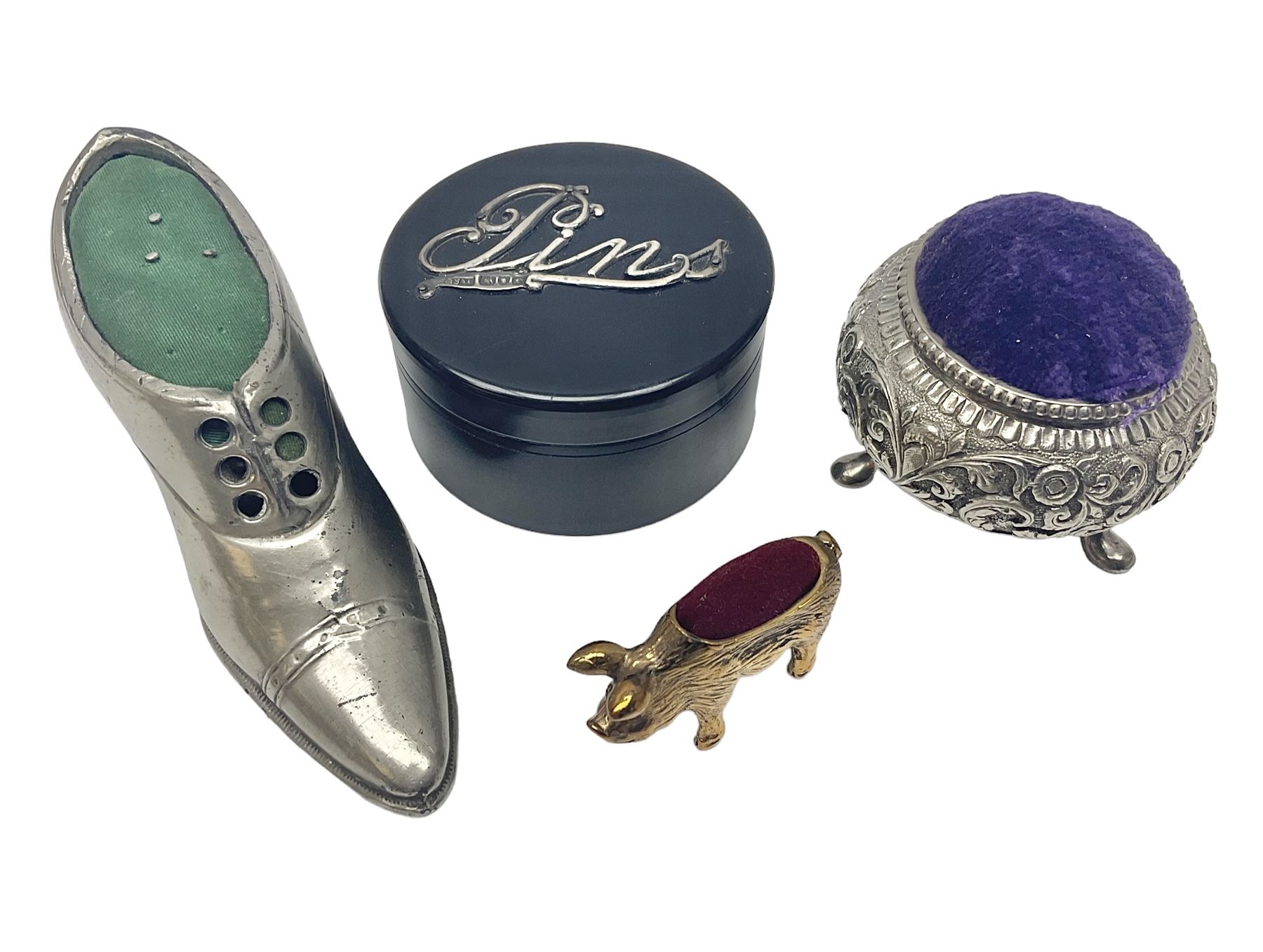 Three pin cushions, comprising gilt metal pig, silver plate shoe, circular pin cushion on threel feet, together with black pin holder with silver writing to the lid 