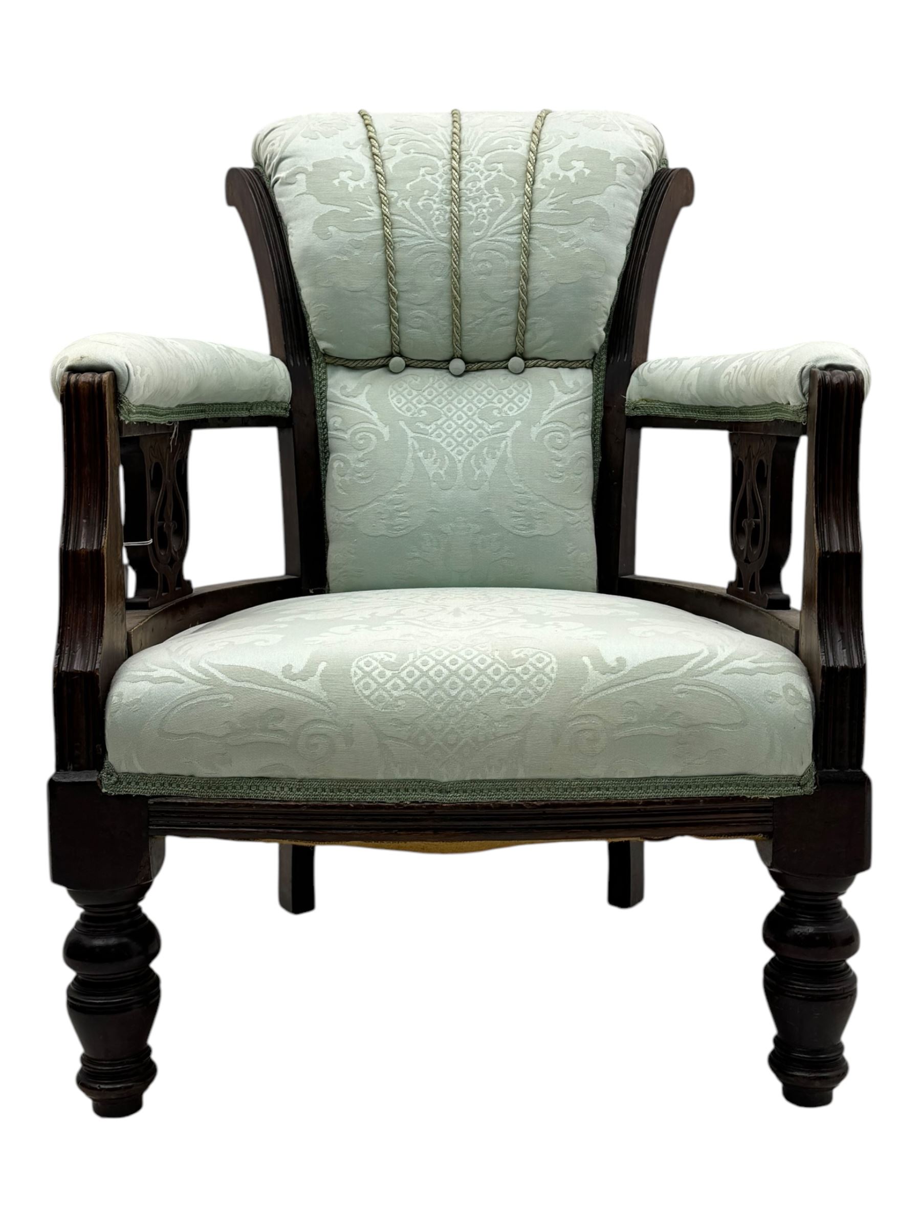 Late Victorian carved mahogany open armchair, the shaped back with fluted upholstered panel and padded arms above pierced scroll carved side supports, over a broad upholstered seat, on turned front supports